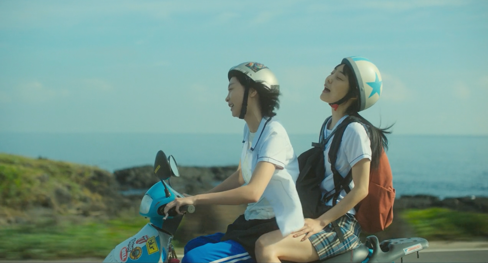 A screenshot from the film Soulmate (2023). It features actresses Kim Da-mi and Jeon So-nee, who are singing and riding a scooter together, a view of the sky, ocean and trees behind them.
