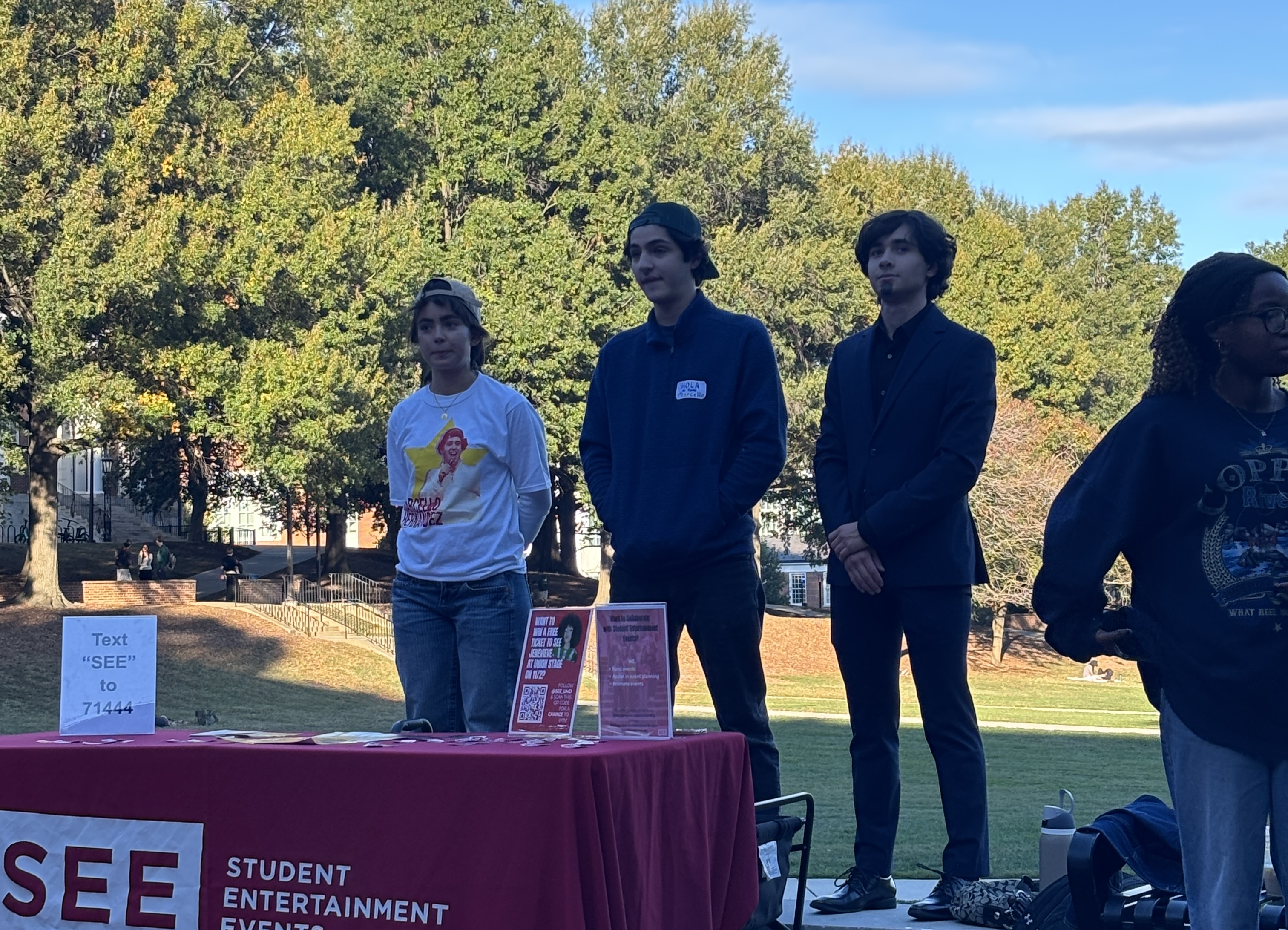 Three students stand next to a table with a \"SEE\" sign, at a student event outdoors. One man wears a white long-sleeve shirt with a graphic, while the other two wear darker attire. They are facing the school library and an audience.