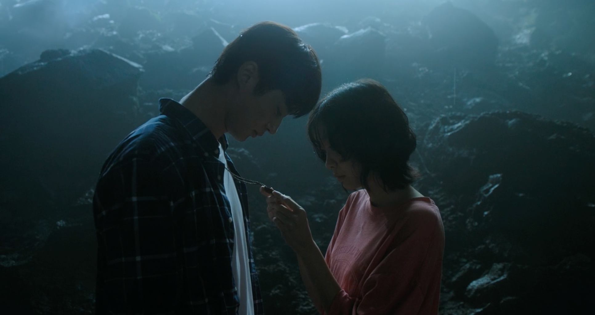 A screenshot from the film Soulmate (2023). It features actress Kim Da-mi and actor Byron Woo-seok, who are standing in a dark cave together, while she holds his necklace and peers at it.