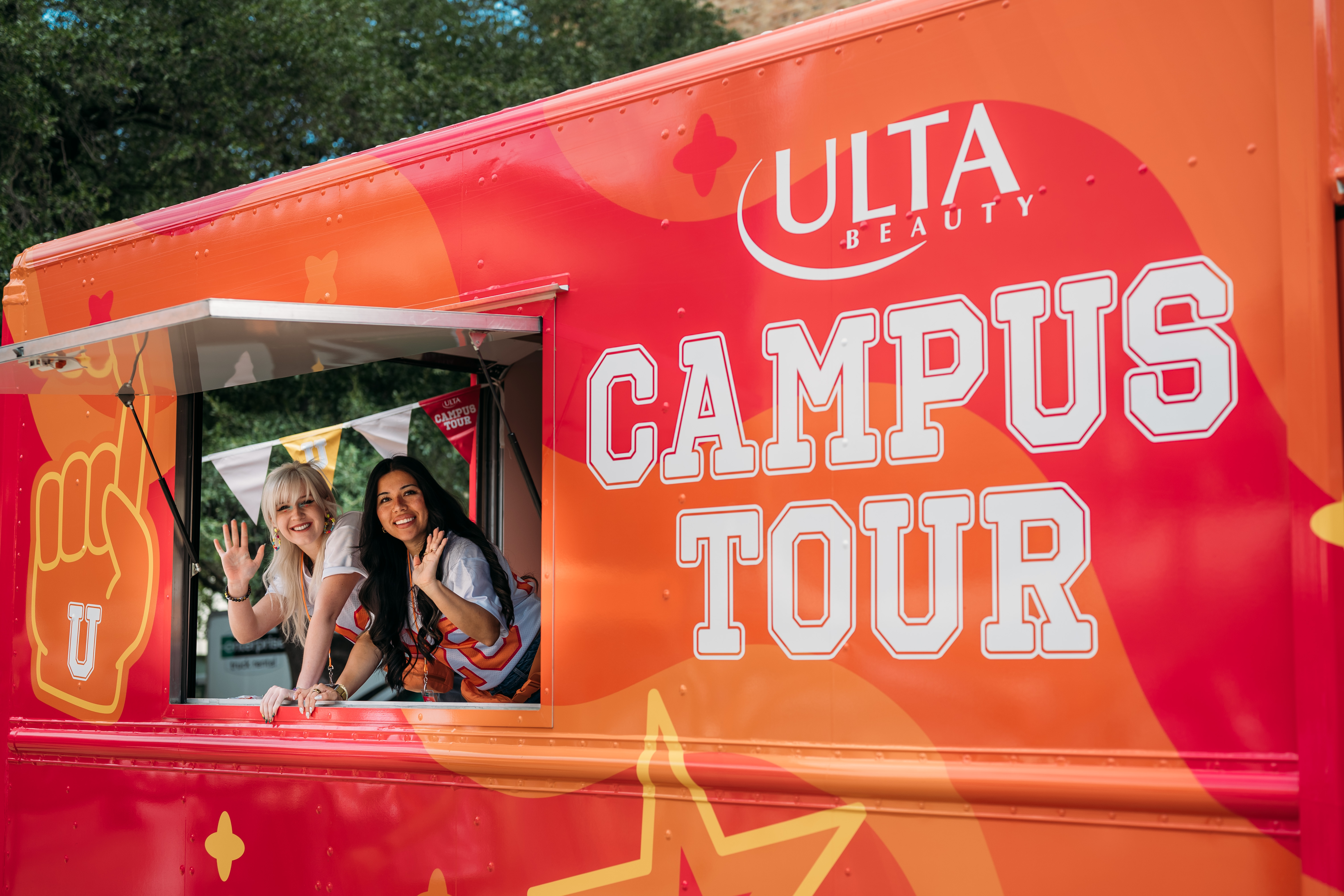 Bright orange Ulta Beauty Campus Tour truck with large lettering and an open side window, where two smiling women wave cheerfully from inside.