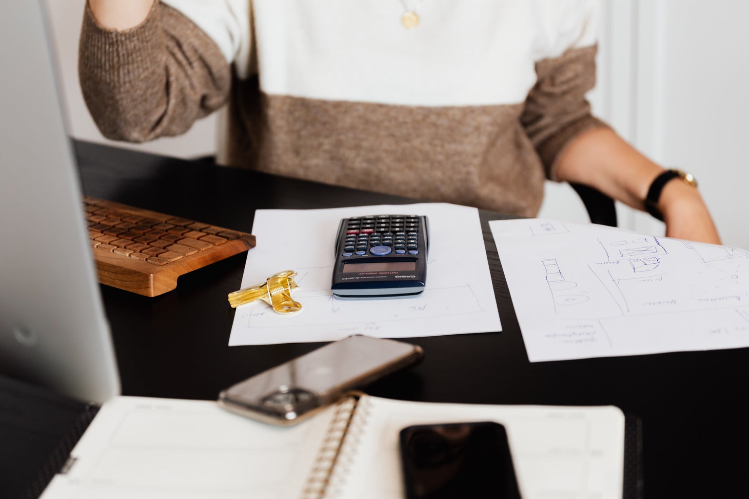 Person in a brown and white sweater sitting in front of computer with a calculator and papers in front of them.