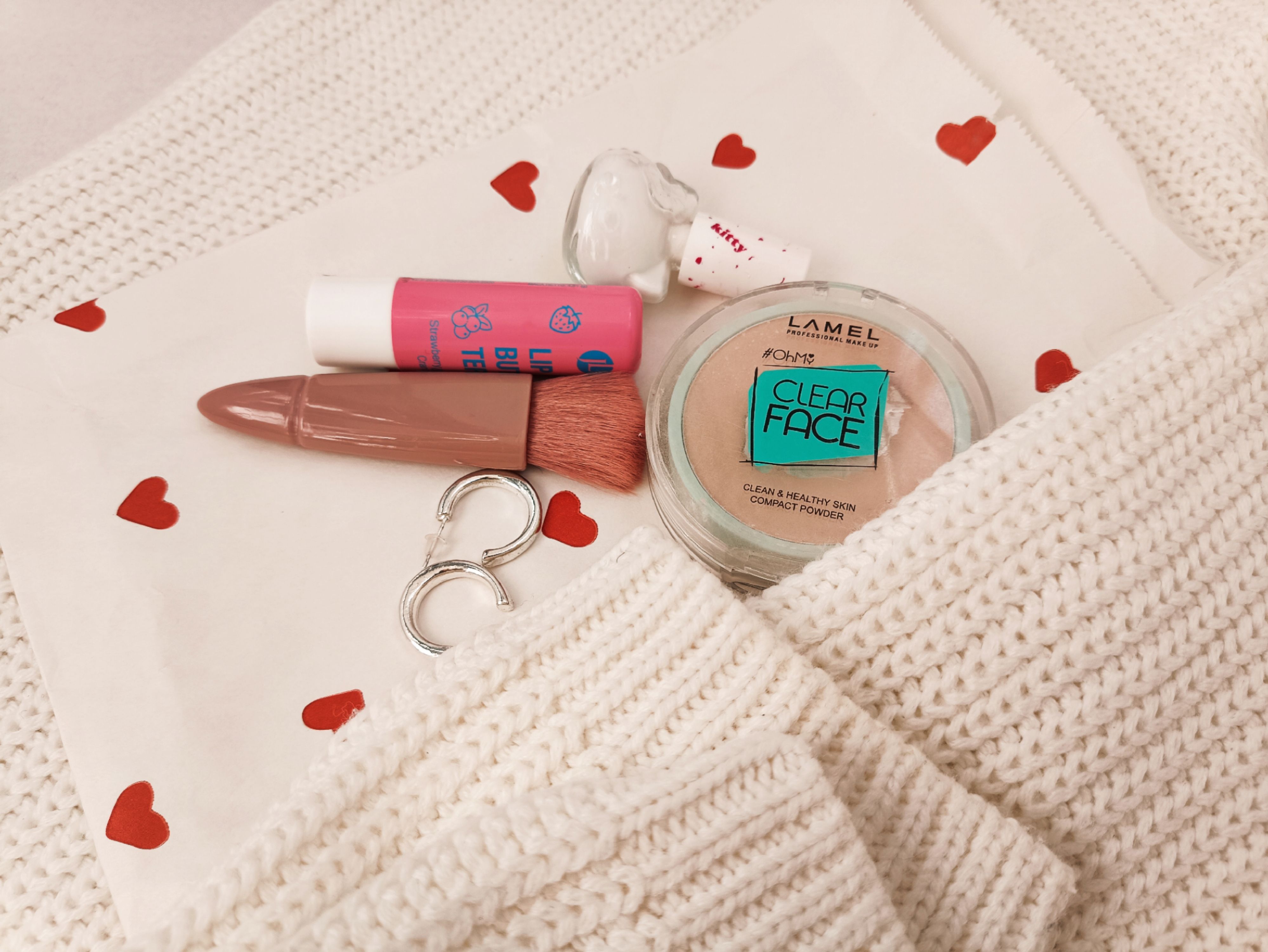 Stylish flat lay featuring makeup items, white knit, and heart-patterned cloth for beauty inspiration