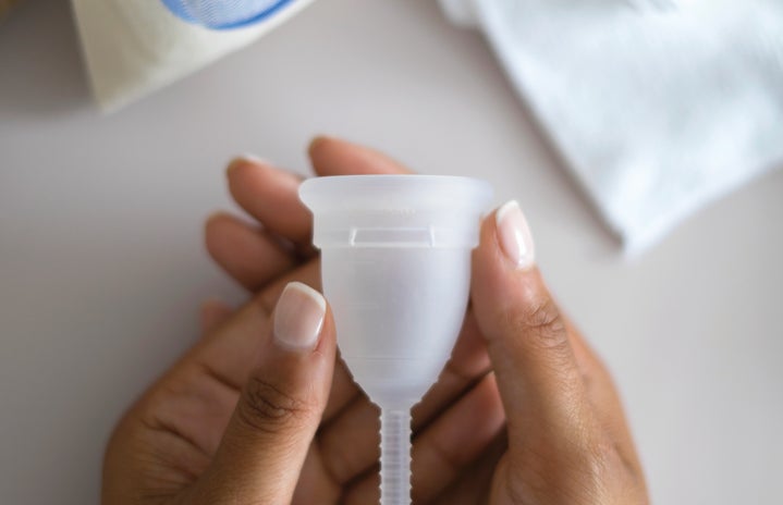 A persons hand holding a clear menstrual cup with a white cloth a white cloth pouch in the background