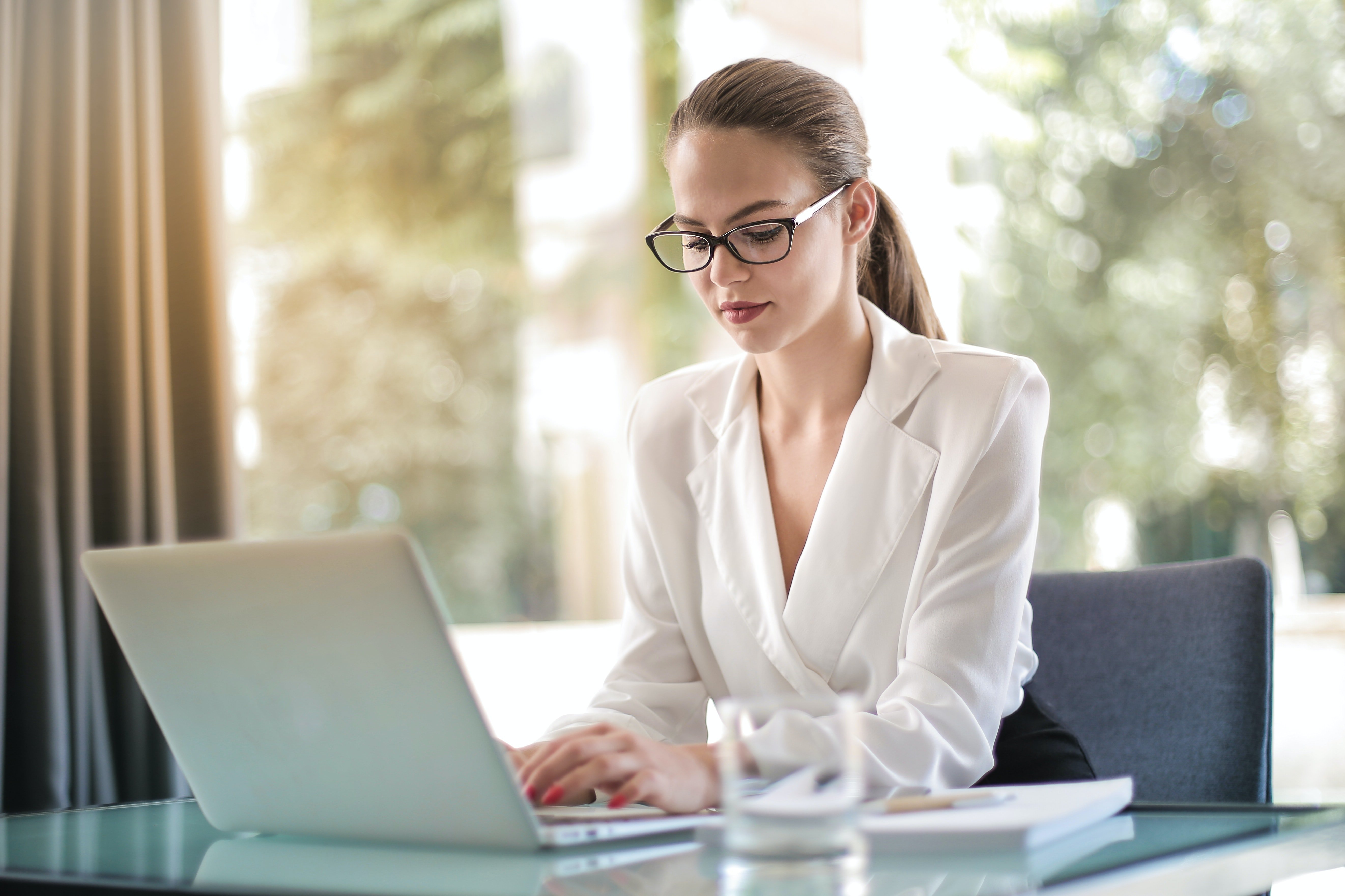 Woman with glasses and hair up sits in front of a window with her laptop in front of her.