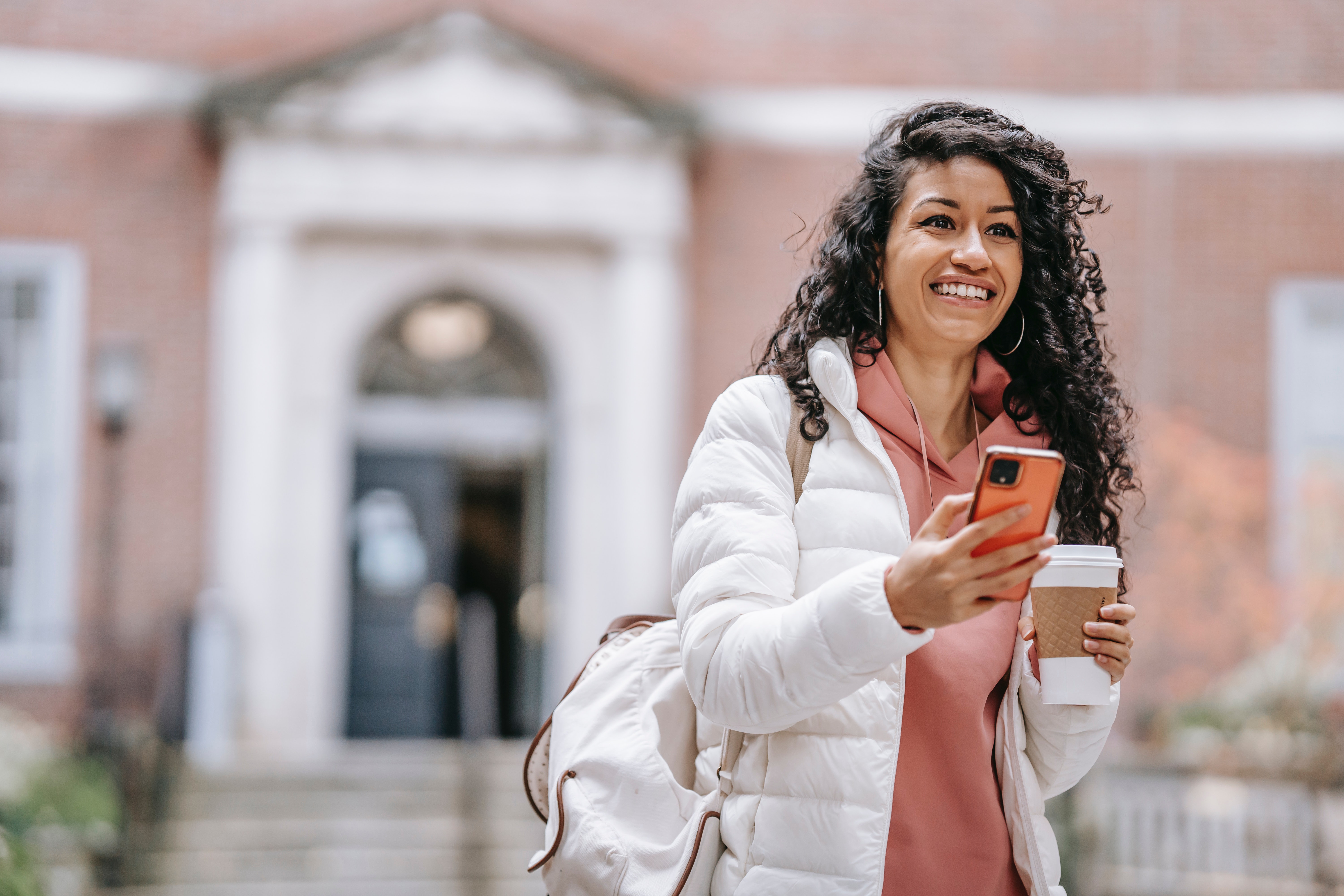 Woman smiles, holds a coffee and her phone, and leaves a building.