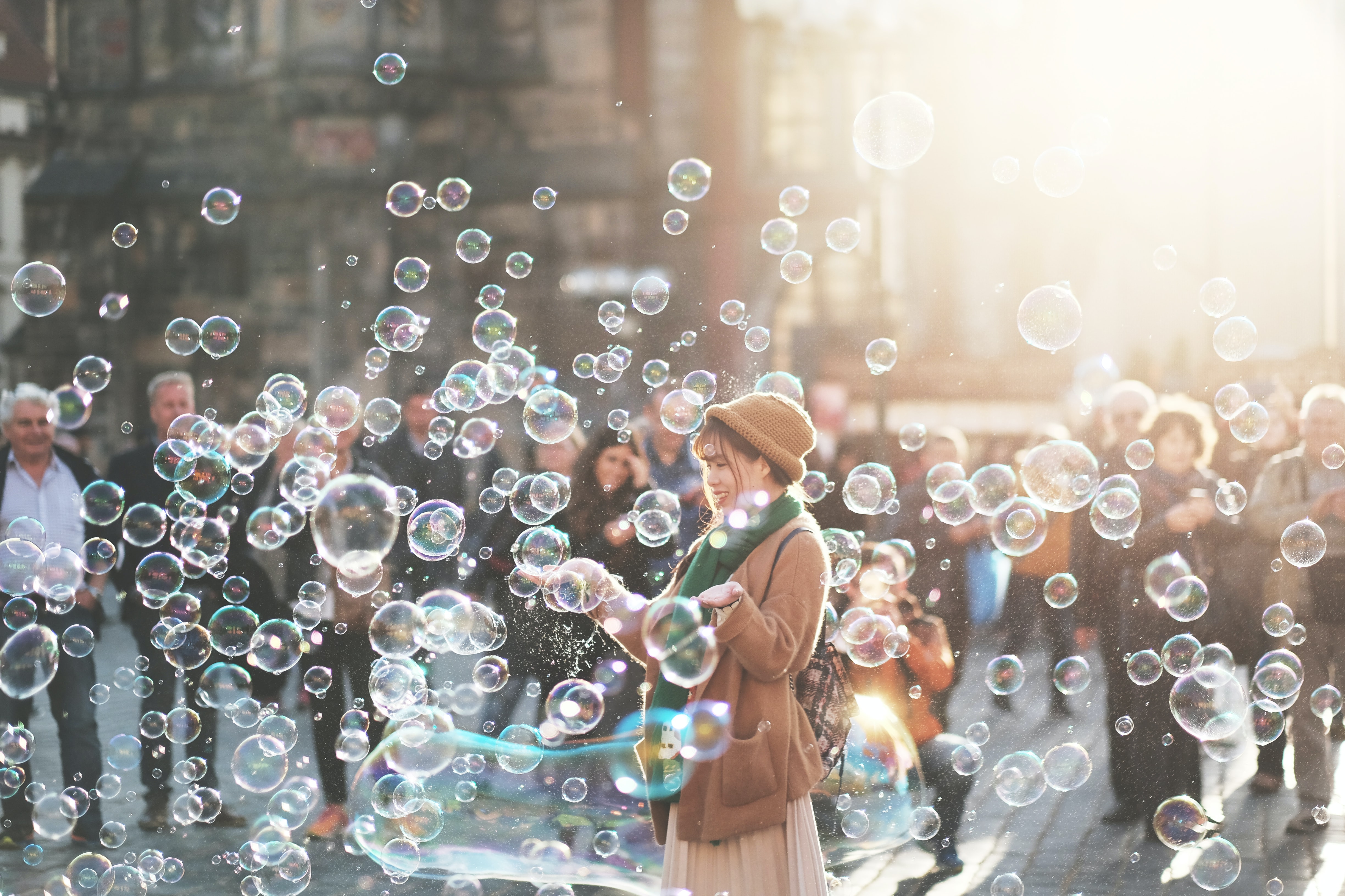 Woman outdoors in a beige coat and hat playing with bubbles, a crowd of folks in the background