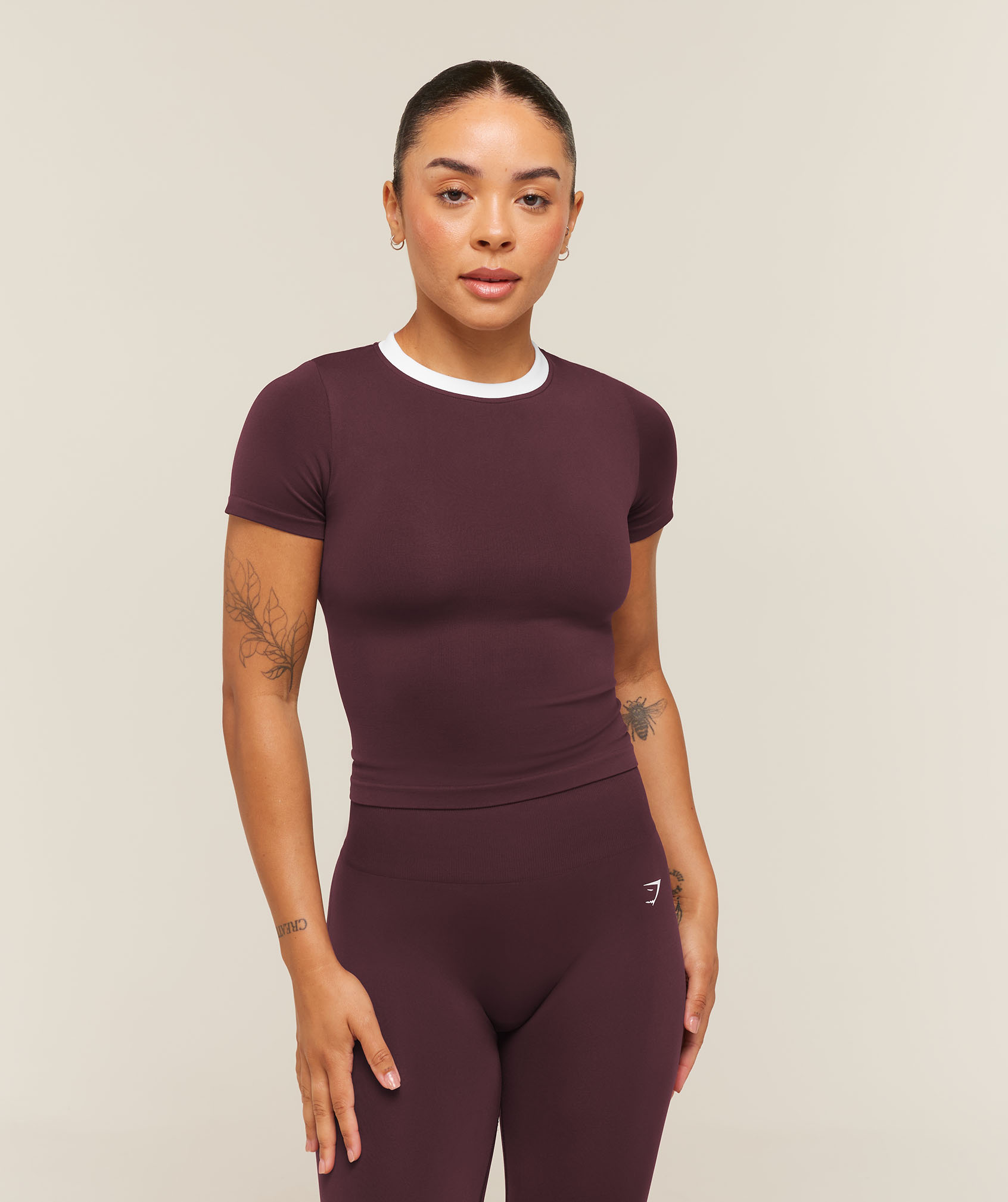 A woman stands against a plain beige studio background, wearing a fitted burgundy athletic top and leggings with white trim, looking calmly at the camera.