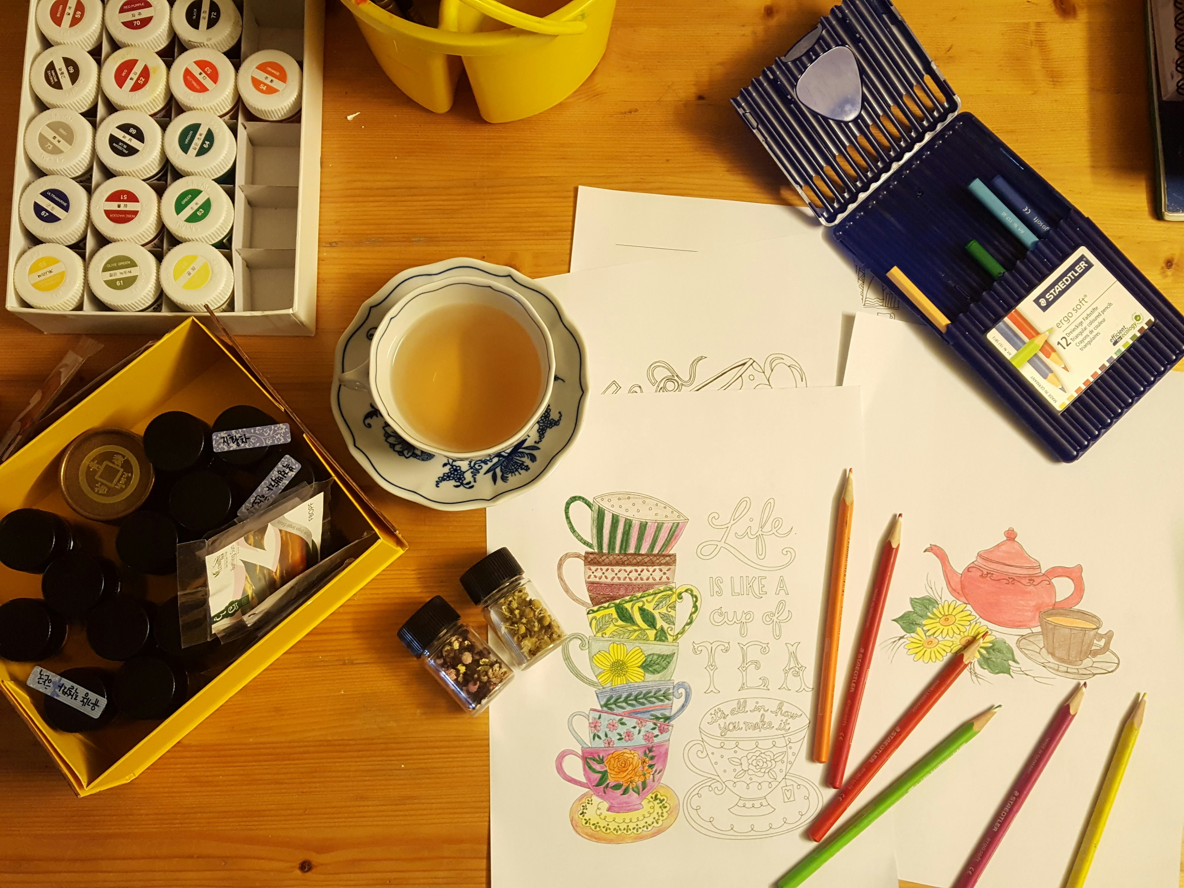 Wooden table with paints, tea cup, coloring pencils, and varies papers with drawings