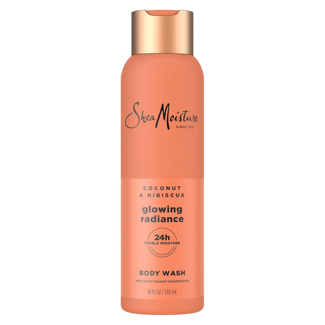 A bottle of Shea Moisture body wash labeled Coconut & Hibiscus, Glowing Radiance. The text highlights 24-hour visible moisture and plant-based moisturizers. The bottle is orange with a copper-colored cap and contains 18 fl oz (532 ml).