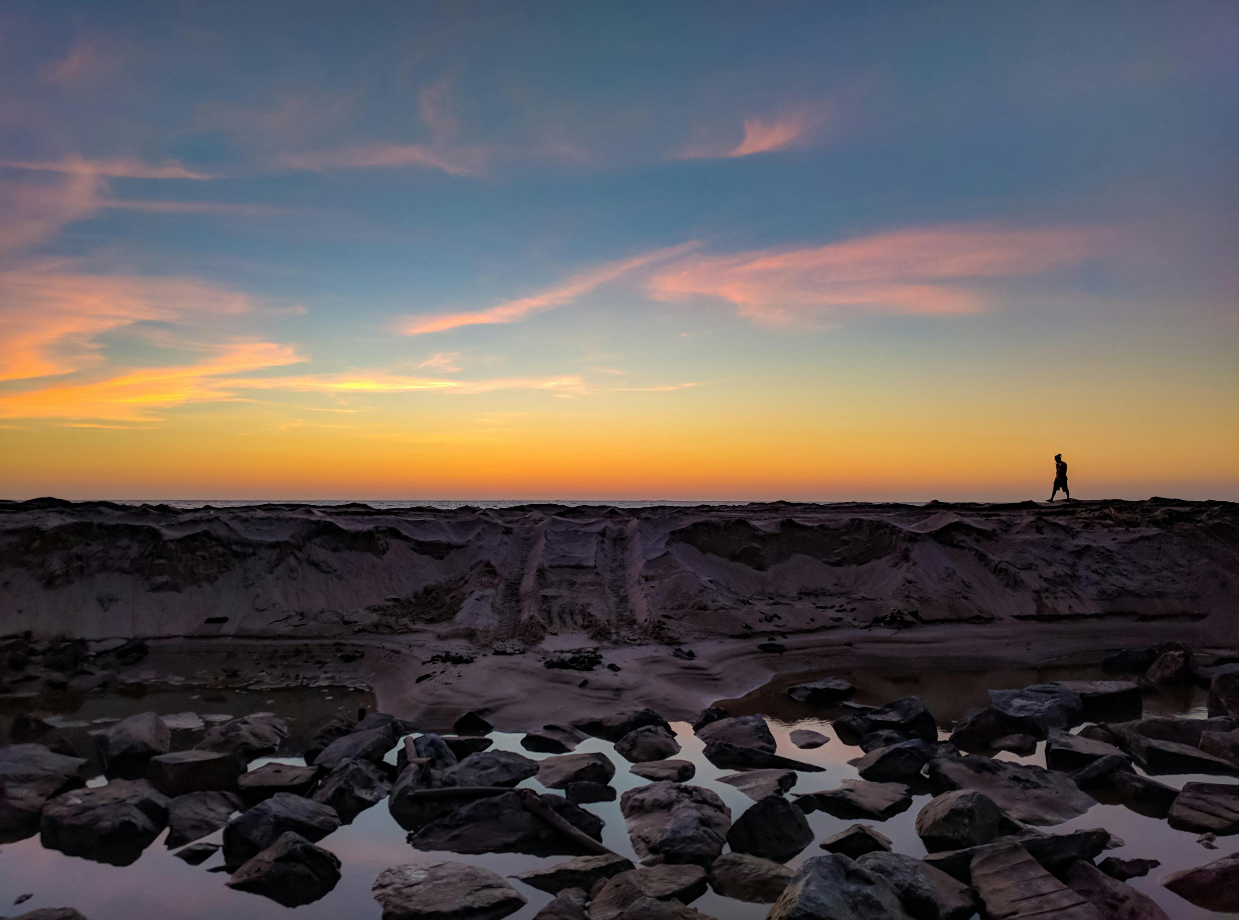 Silhouette of person walking on rocks, water at sunset