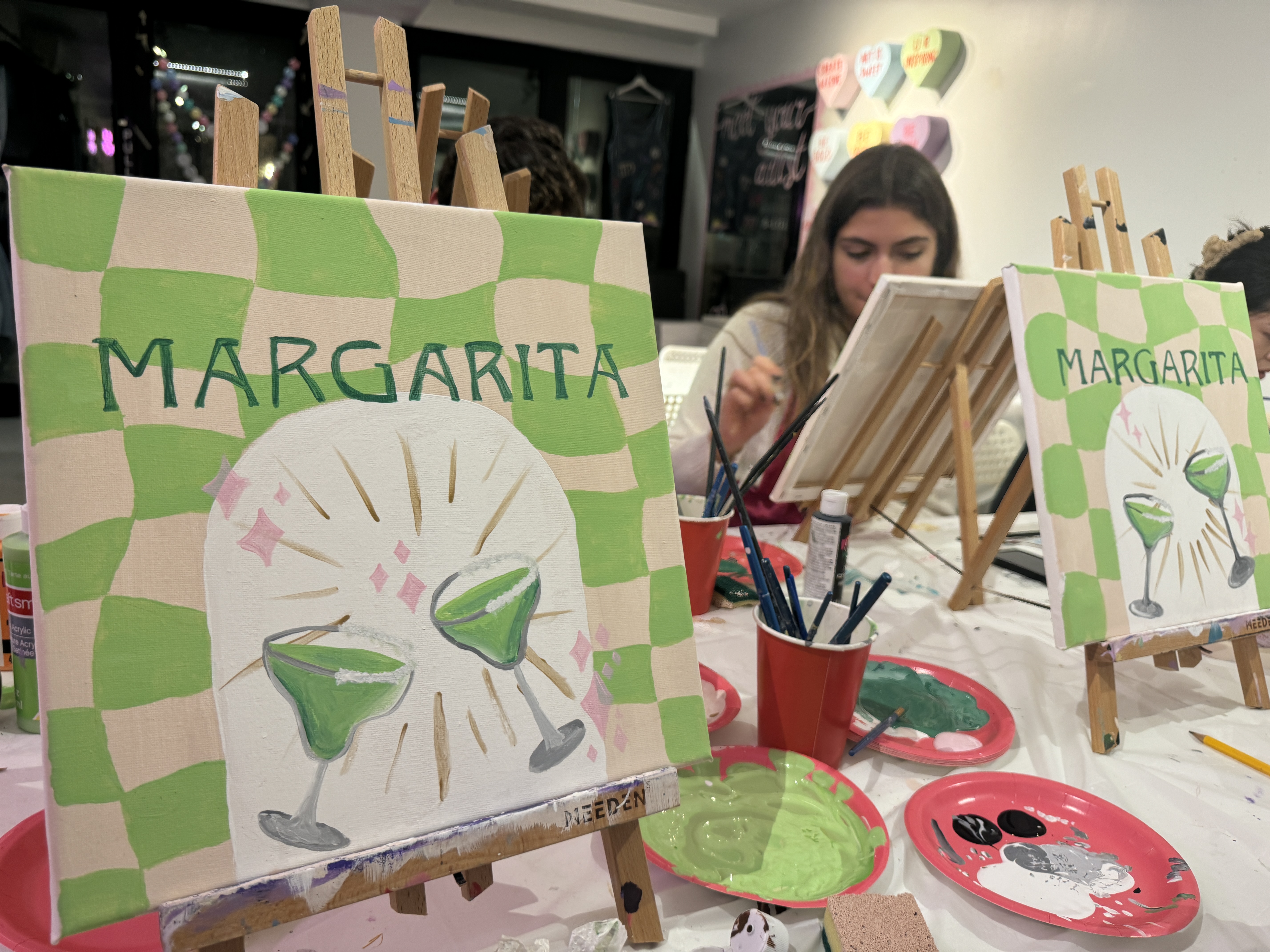 Two easels holding up paintings of green margaritas with green and beige checkered backgrounds, surrounded by art supplies on a table.