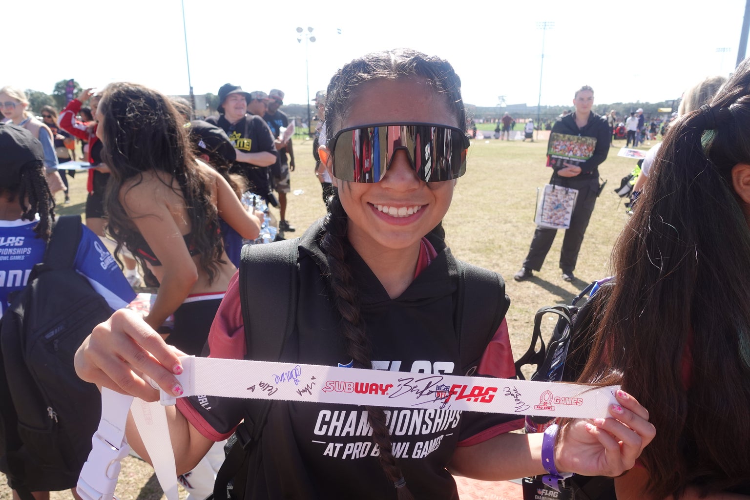 A young female flag football player wearing glasses and proudly holding up her flag football belt that has been signed by the NFL cheerleaders