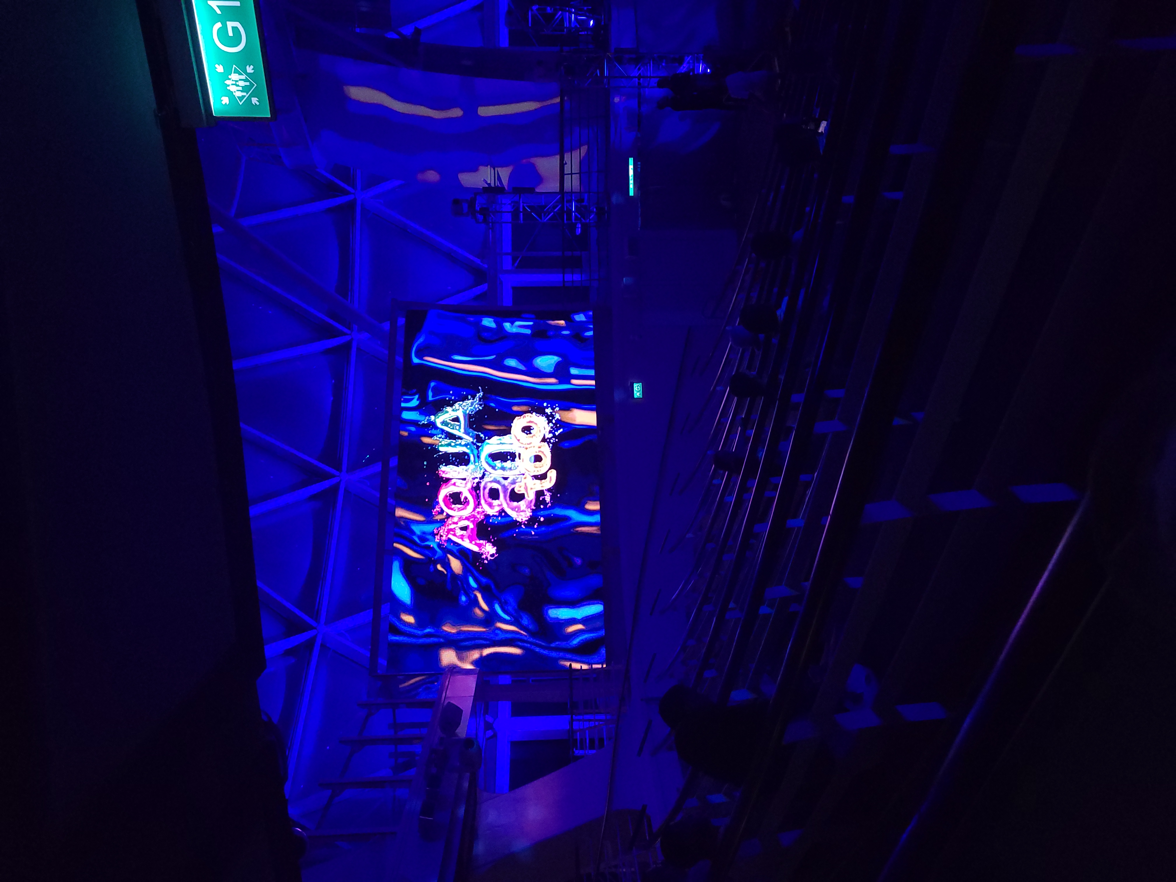 A dark lit picture of the outdoor aquatheatre screens featuring the name of the show \'Aqua80too\' in shimmery and water-like bubble font