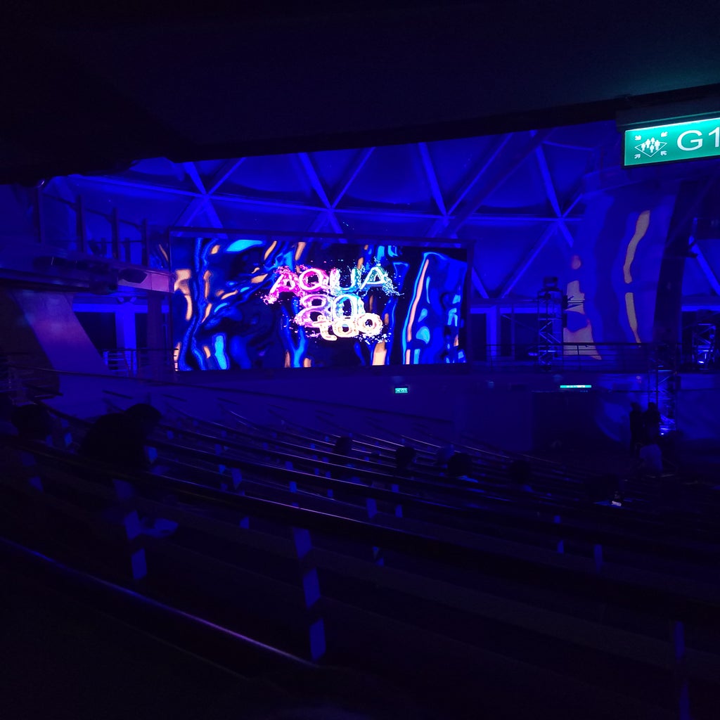 A dark lit picture of the outdoor aquatheatre screens featuring the name of the show \'Aqua80too\' in shimmery and water-like bubble font