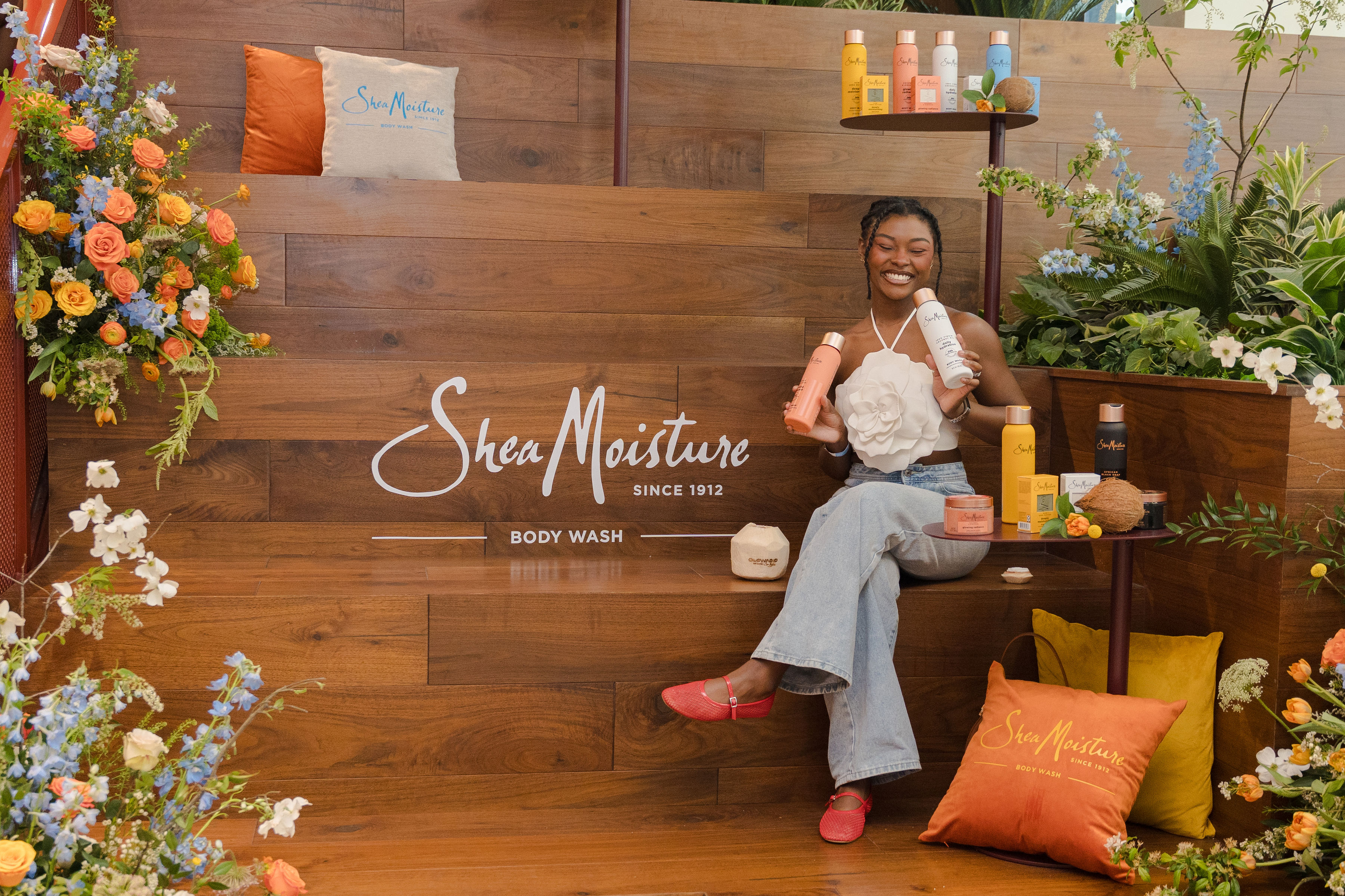 A woman in a white top and jeans sits on a wooden ledge surrounded by colorful flowers and SheaMoisture body wash products. The background has a wooden texture with the SheaMoisture logo. She smiles while holding a bottle.