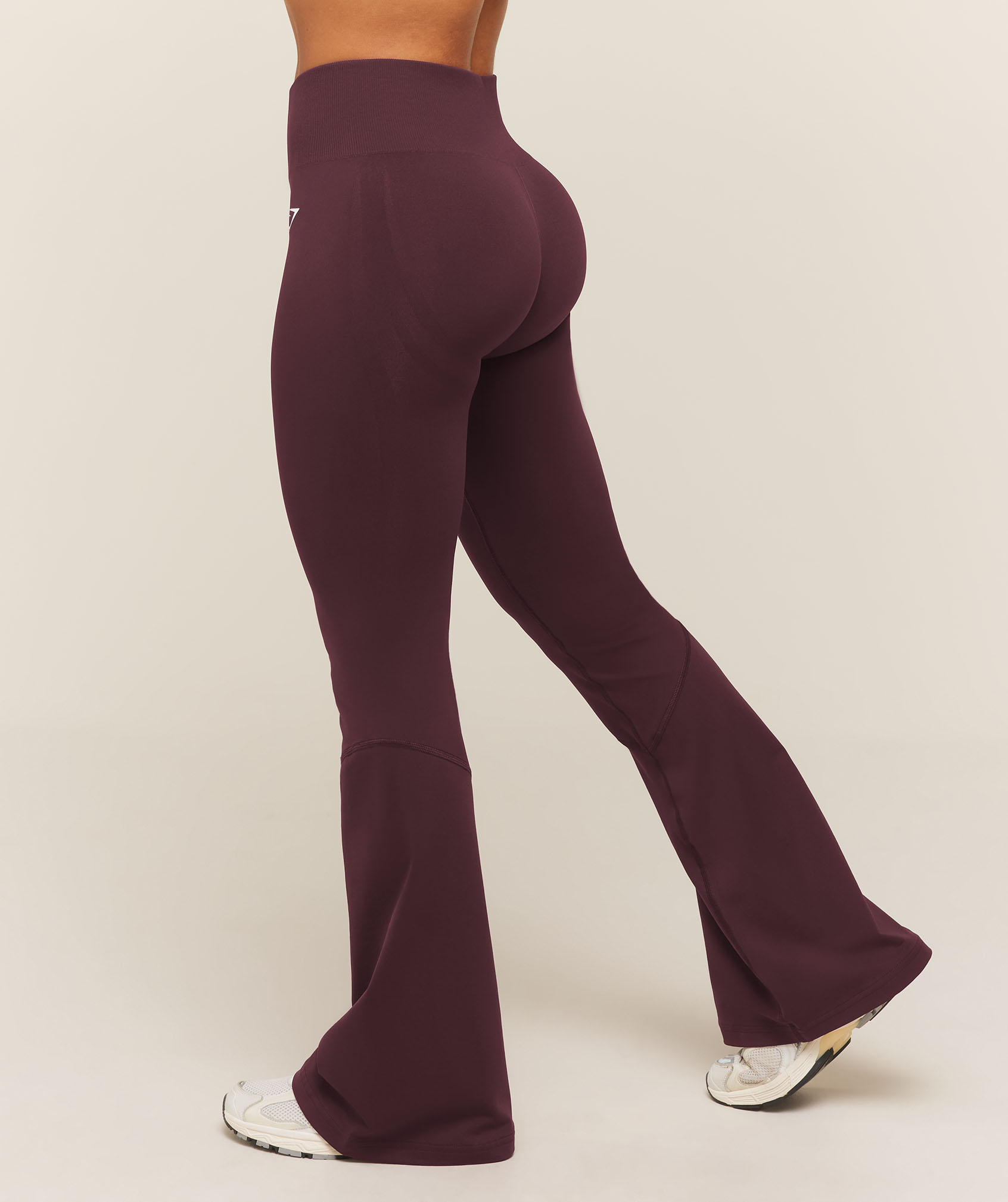 A model poses in plum high-waisted flared leggings and white sneakers against a neutral studio backdrop, highlighting the fit and silhouette.