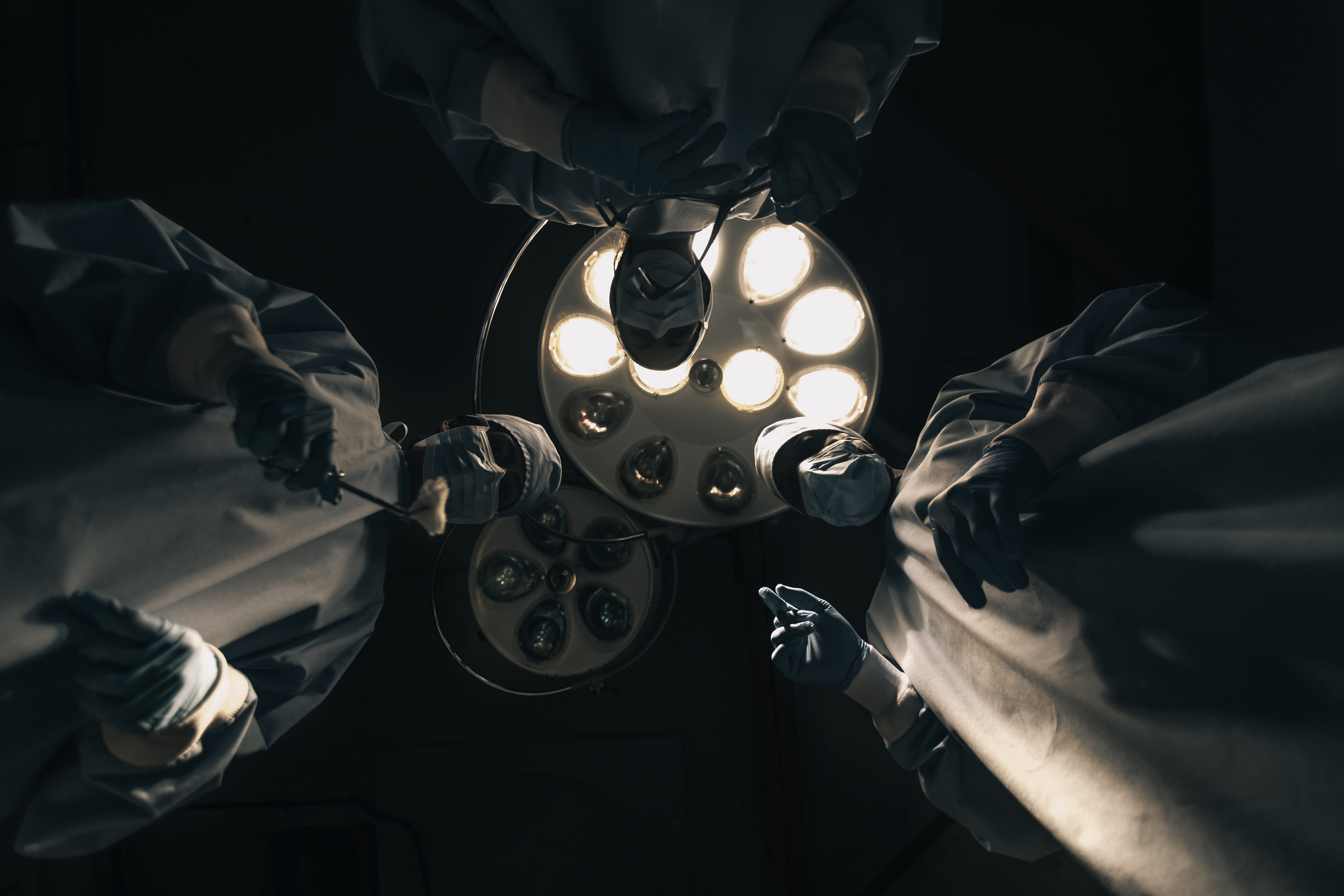 A point of view shot from an operating table staring up at three surgeons backlit by powerful operating lights.