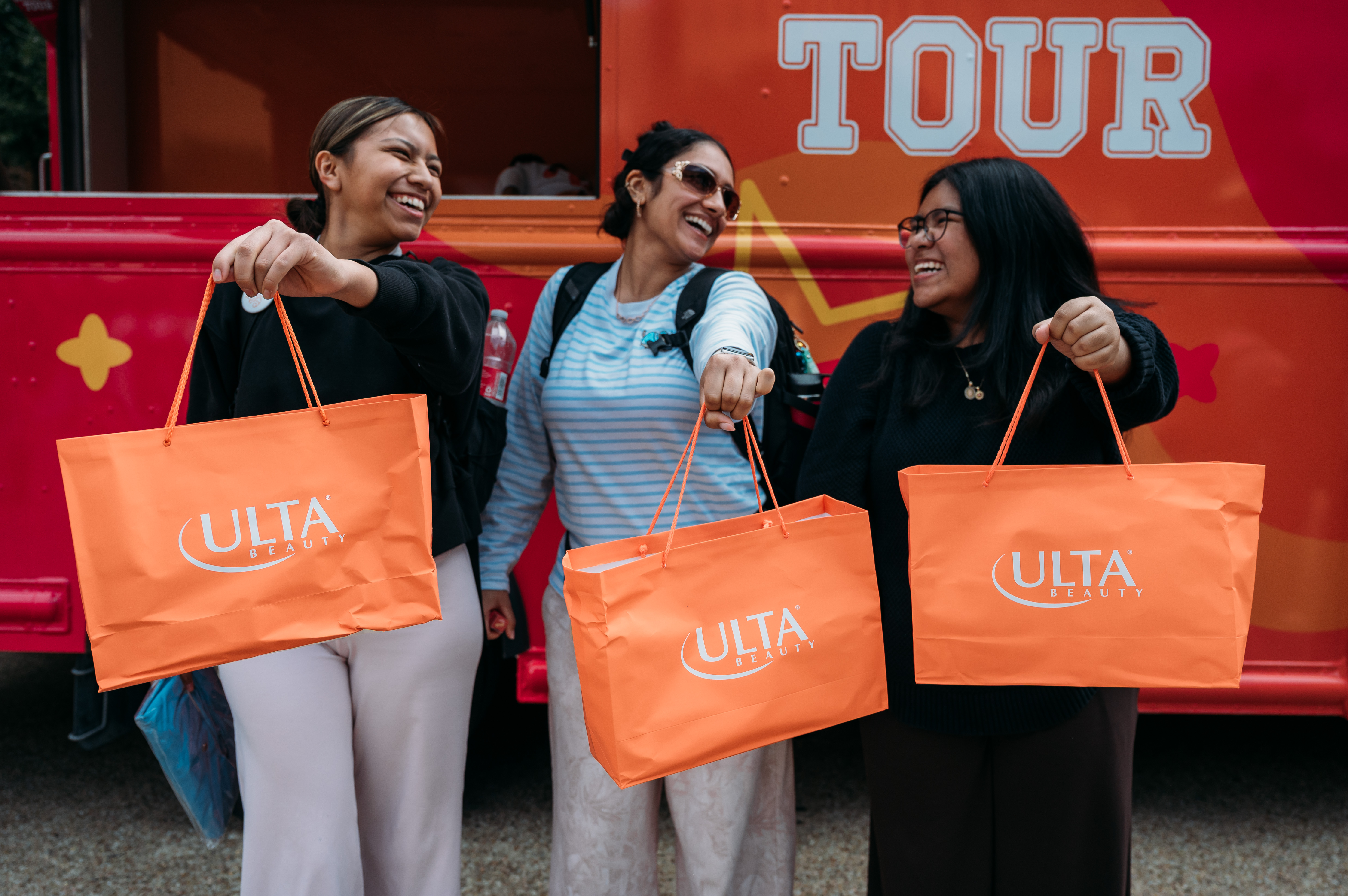 Three smiling women pose with orange Ulta Beauty shopping bags in front of a bright red tour bus, conveying a cheerful, friendly shopping-trip mood.