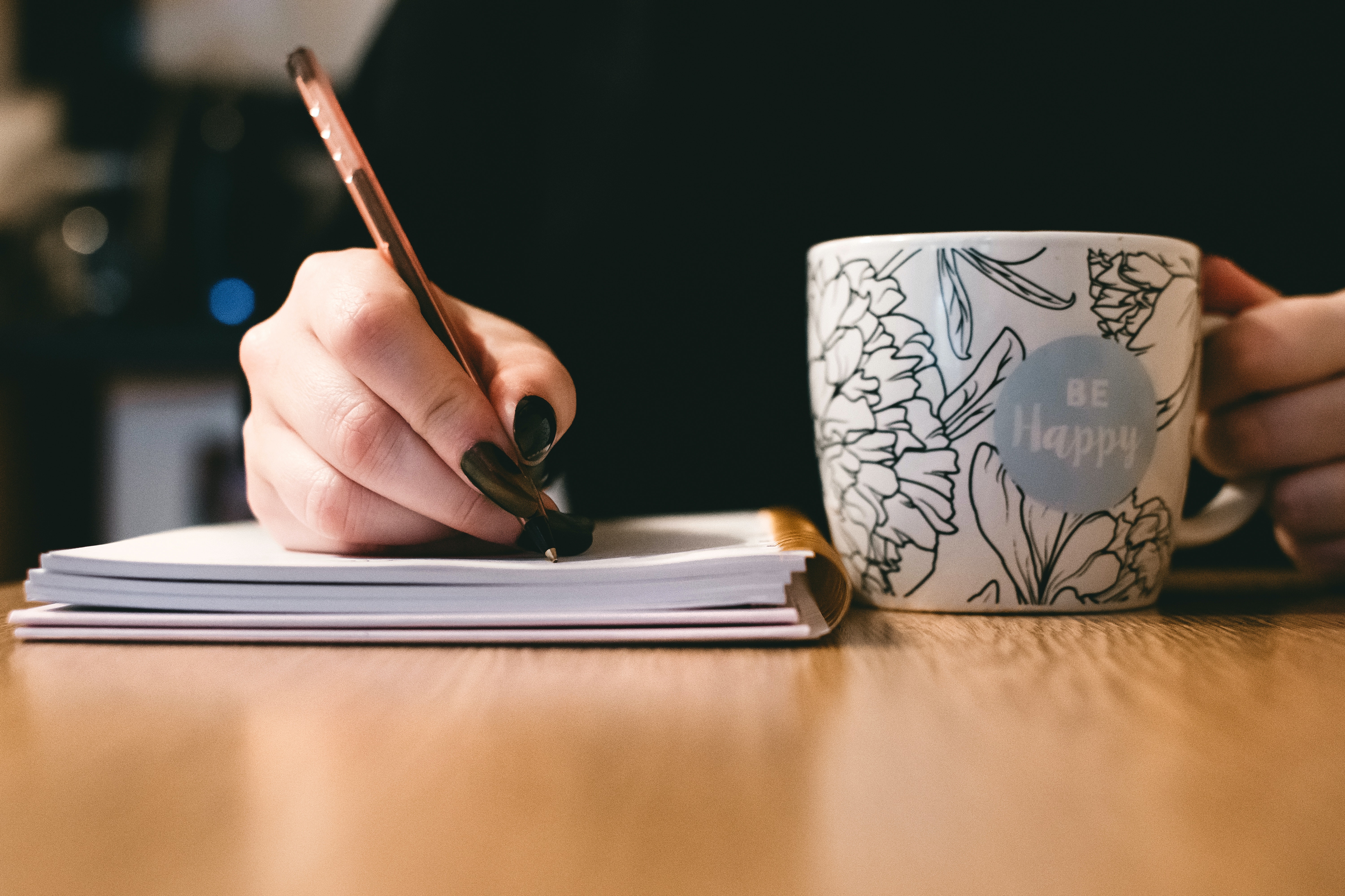 A woman with dark nail polish writers and holds a mug.