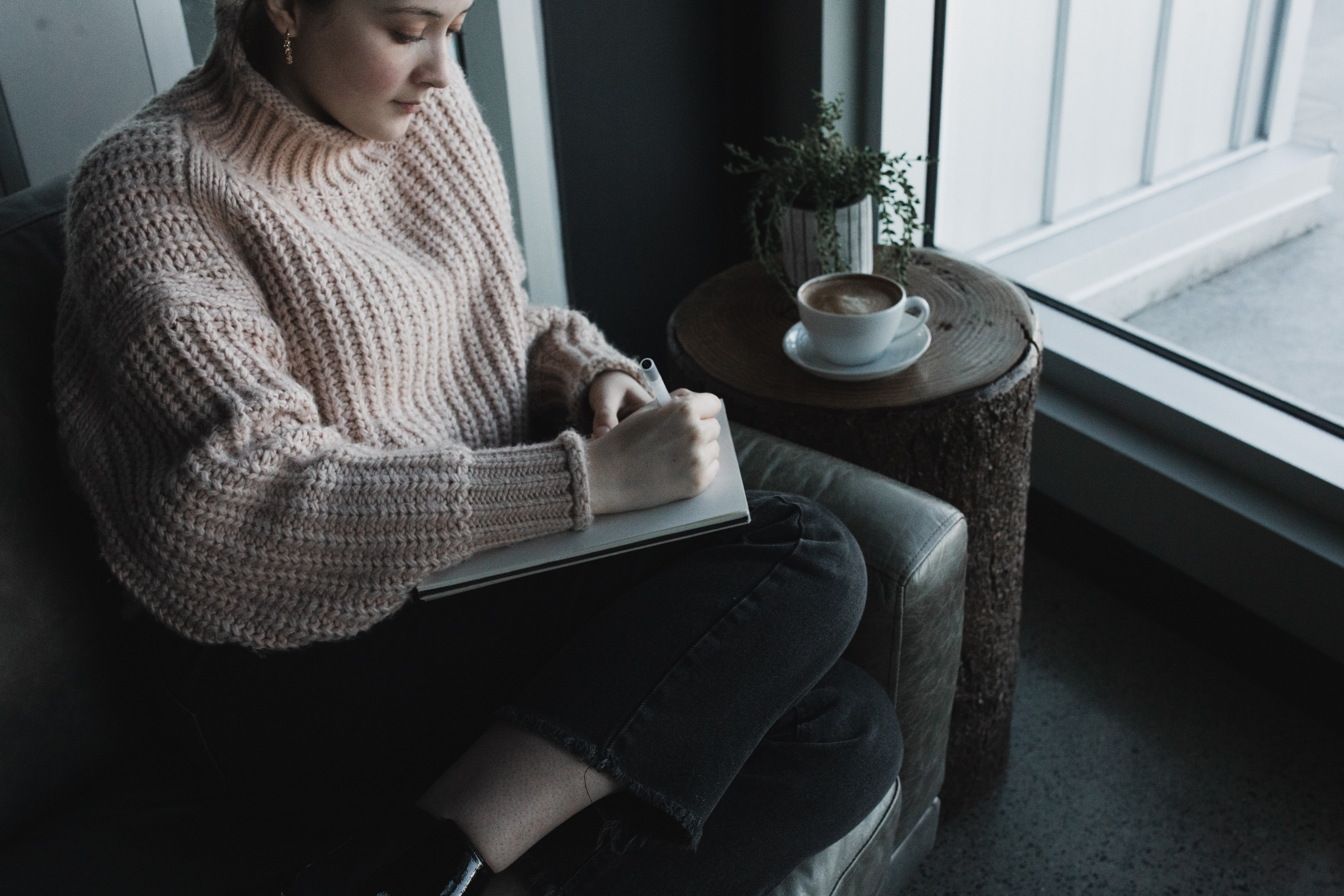 Woman in a light pink sweater sitting beside a window writing in her journal