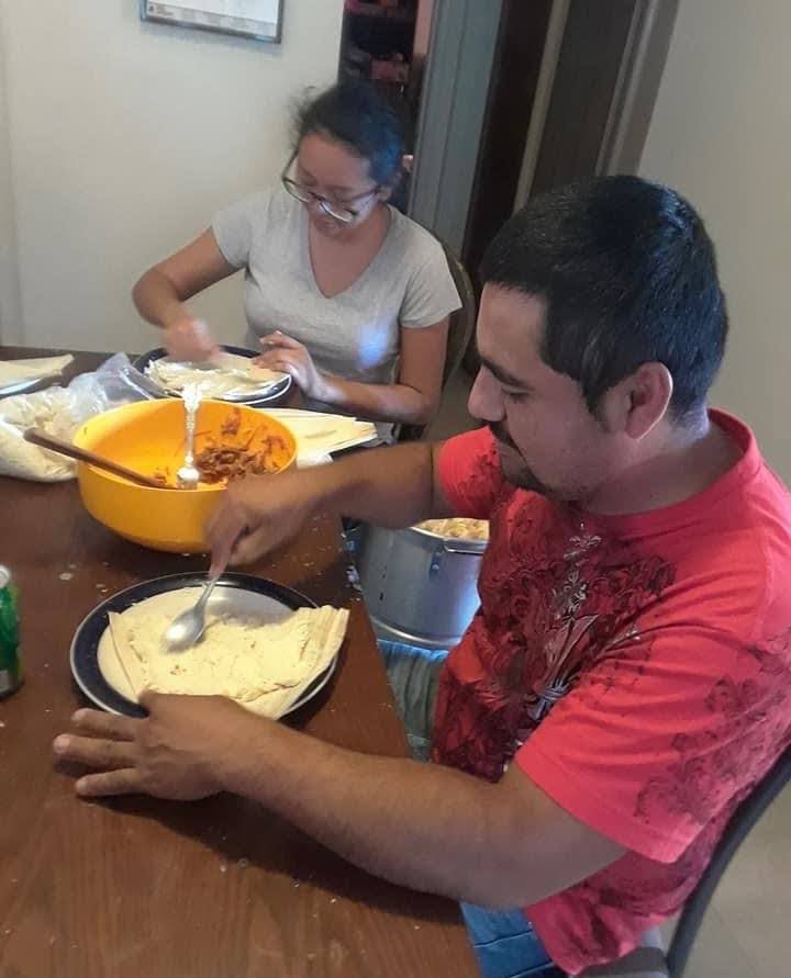 A man and a woman are seated at a table preparing food. The man in a red shirt is spreading masa on corn husks, while the woman in a gray shirt works beside a bowl of filling. The scene conveys a sense of teamwork and focus.