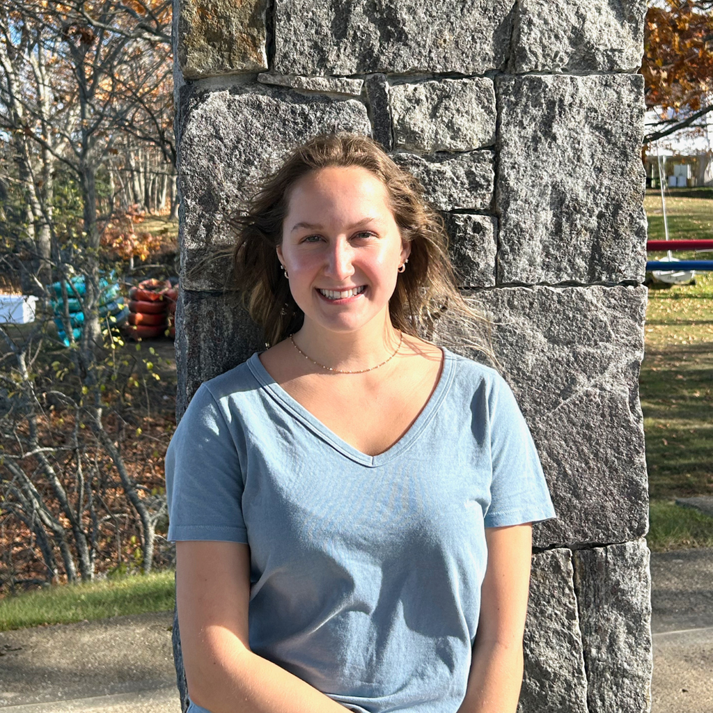 woman with light brown hair standing in front of a stone wall wearing a light blue v-neck shirt