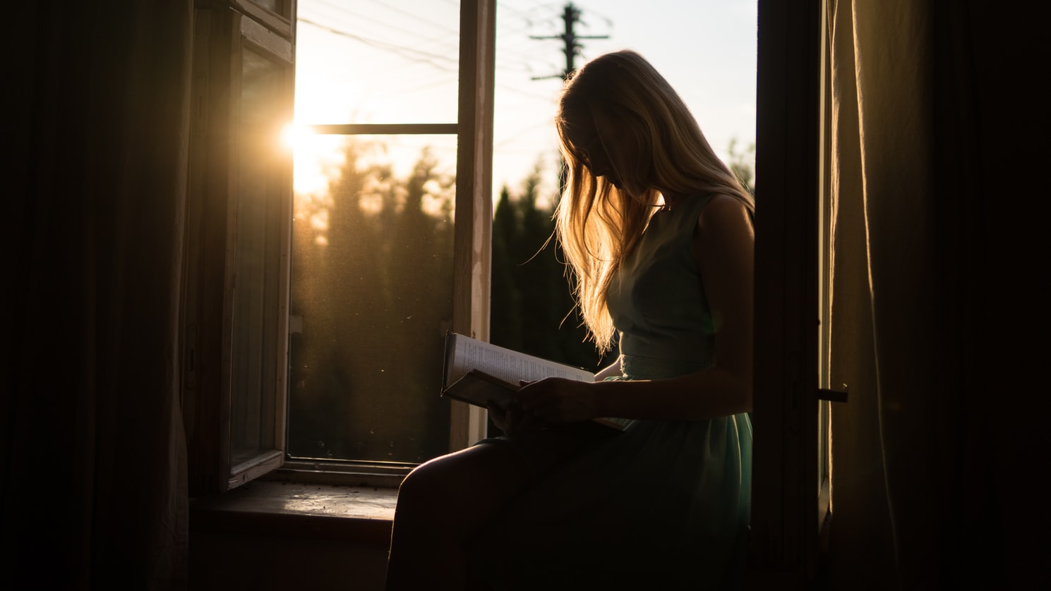 Woman reading on windowsill with sunset in background