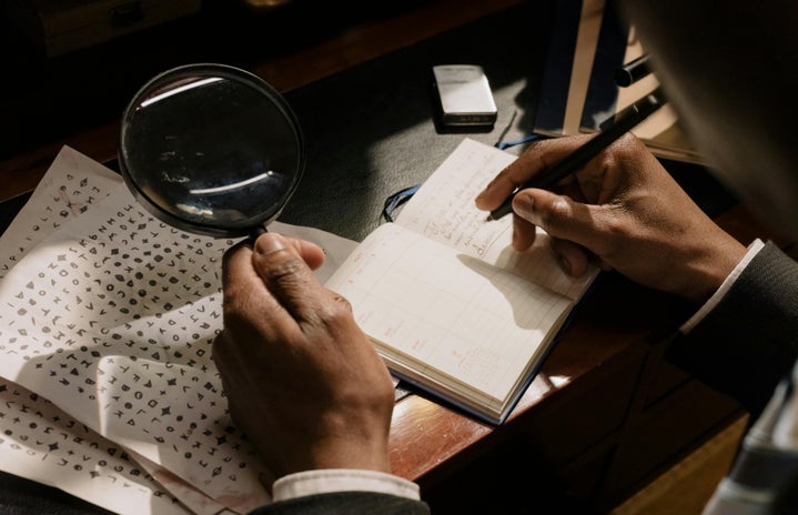 Individual holding a magnifying glass investigating a notebook and an encrypted piece of paper on a table.