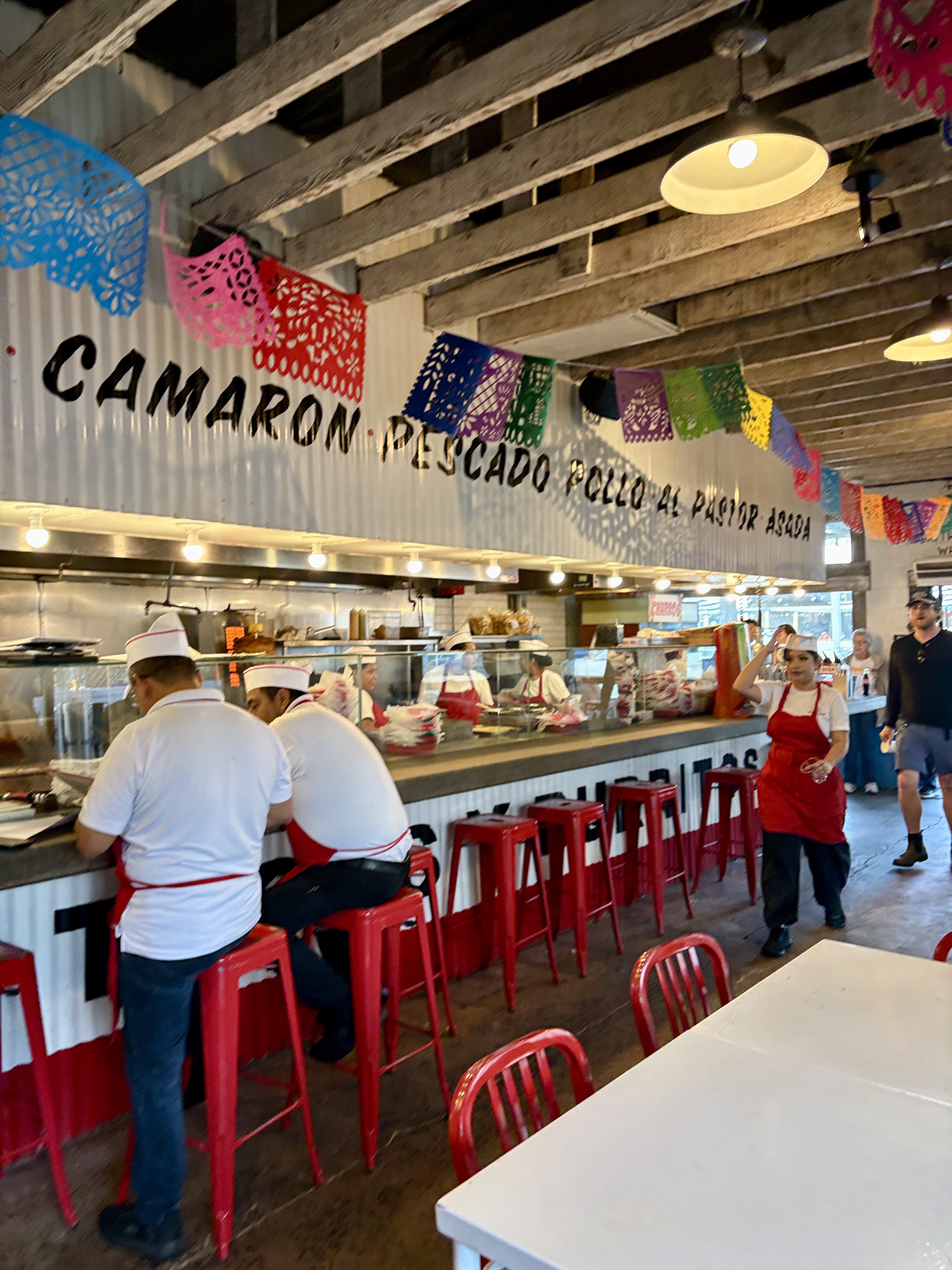 Interior of The Taco Stand showing kitchen staff in red aprons working behind the counter, with menu items painted on the wall above.