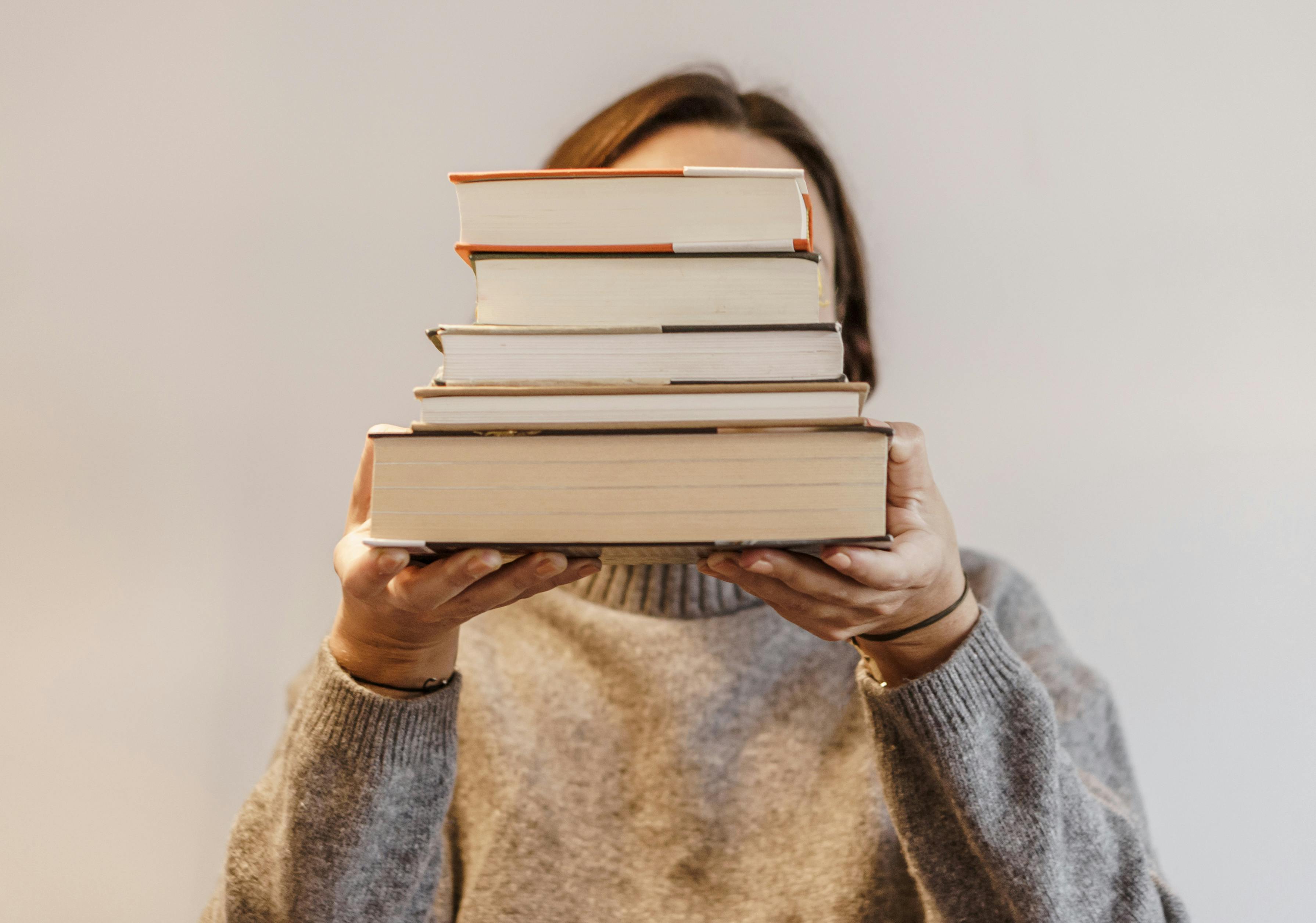 photo of women holding a stack of books on a plain background hiding her face