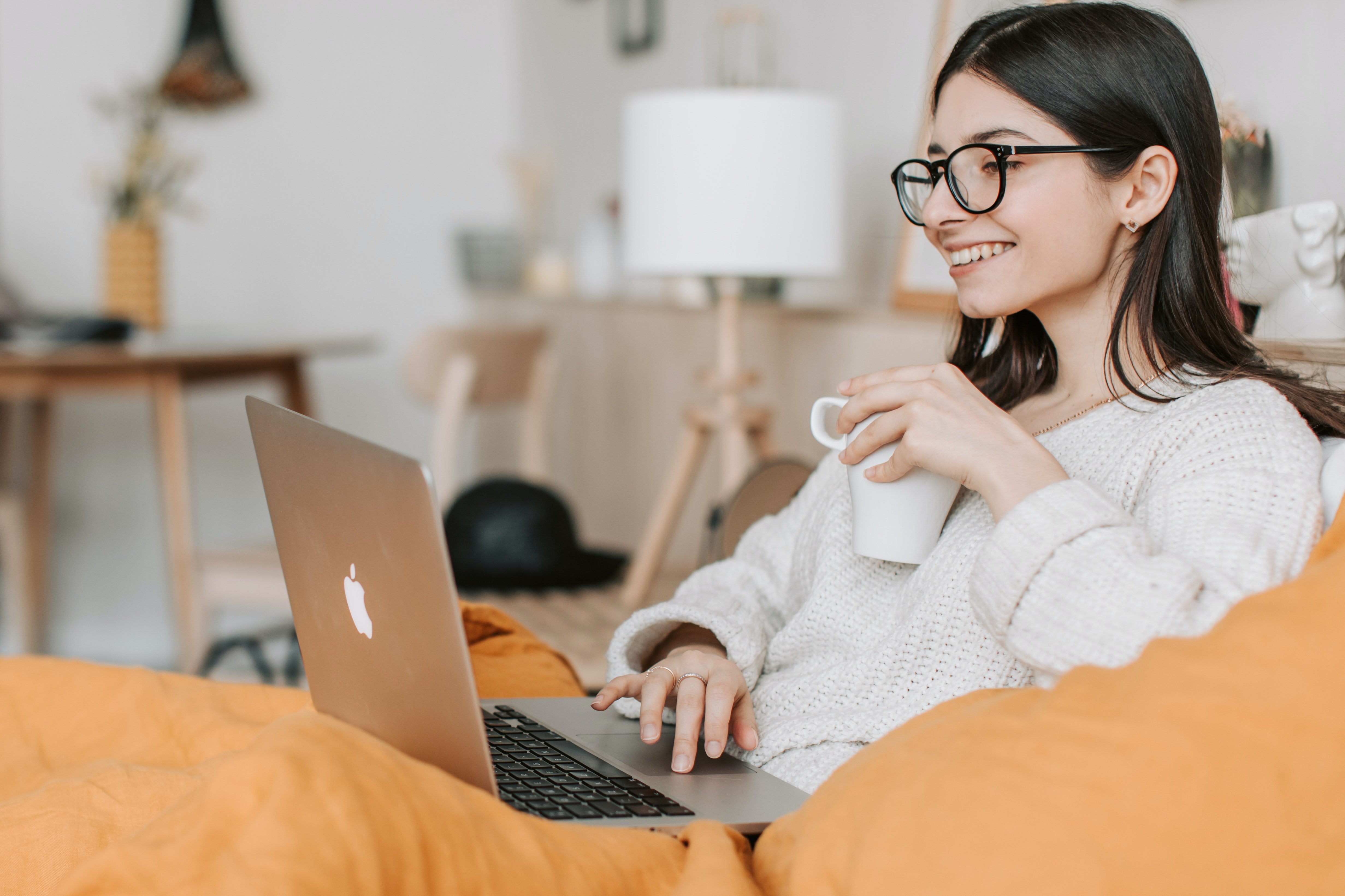 Woman with glasses sitting on a couch with bright orange pillows with a mug in her hand and laptop in front of her.