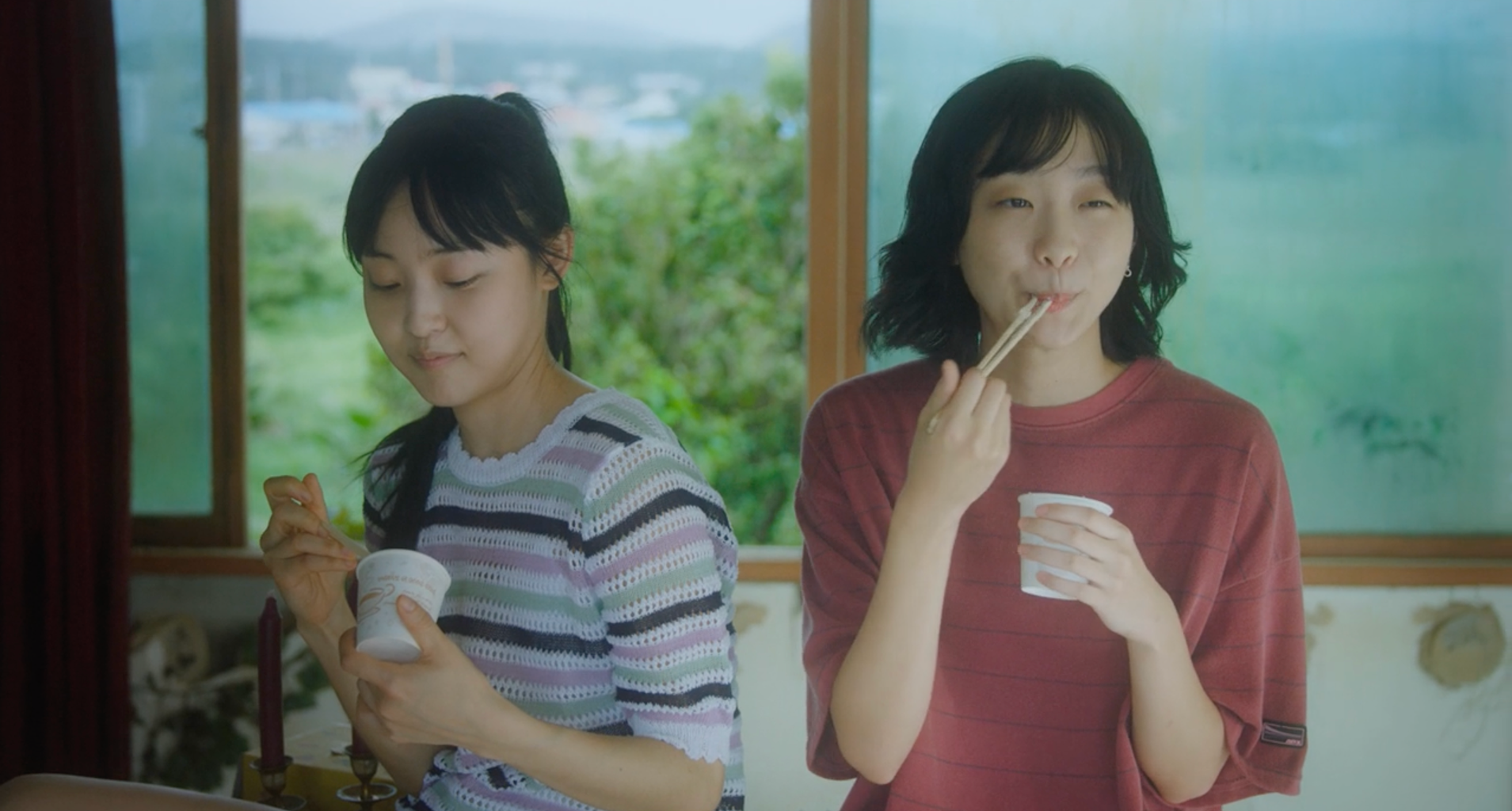 A screenshot from the film Soulmate (2023). It features actresses Kim Da-mi and Jeon So-nee, who are sitting side by side. eating cup noodles, with trees and the sky behind them/