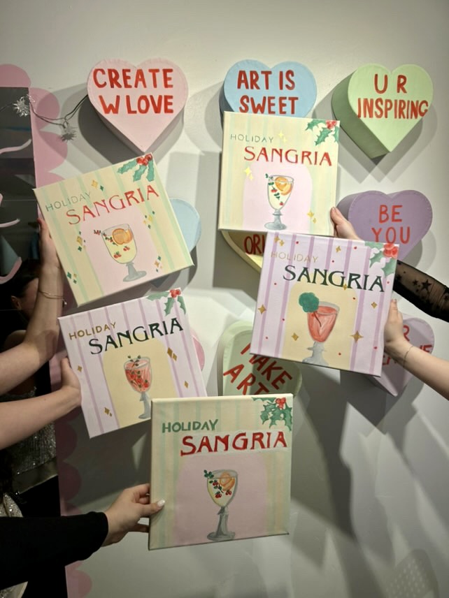 A group of hands holding up paint canvases of holiday-themed sangrias, surrounded by decorative heart-shaped messages behind them on a wall