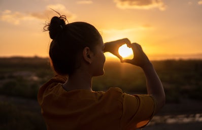 girl making a heart with her hands over a sunset