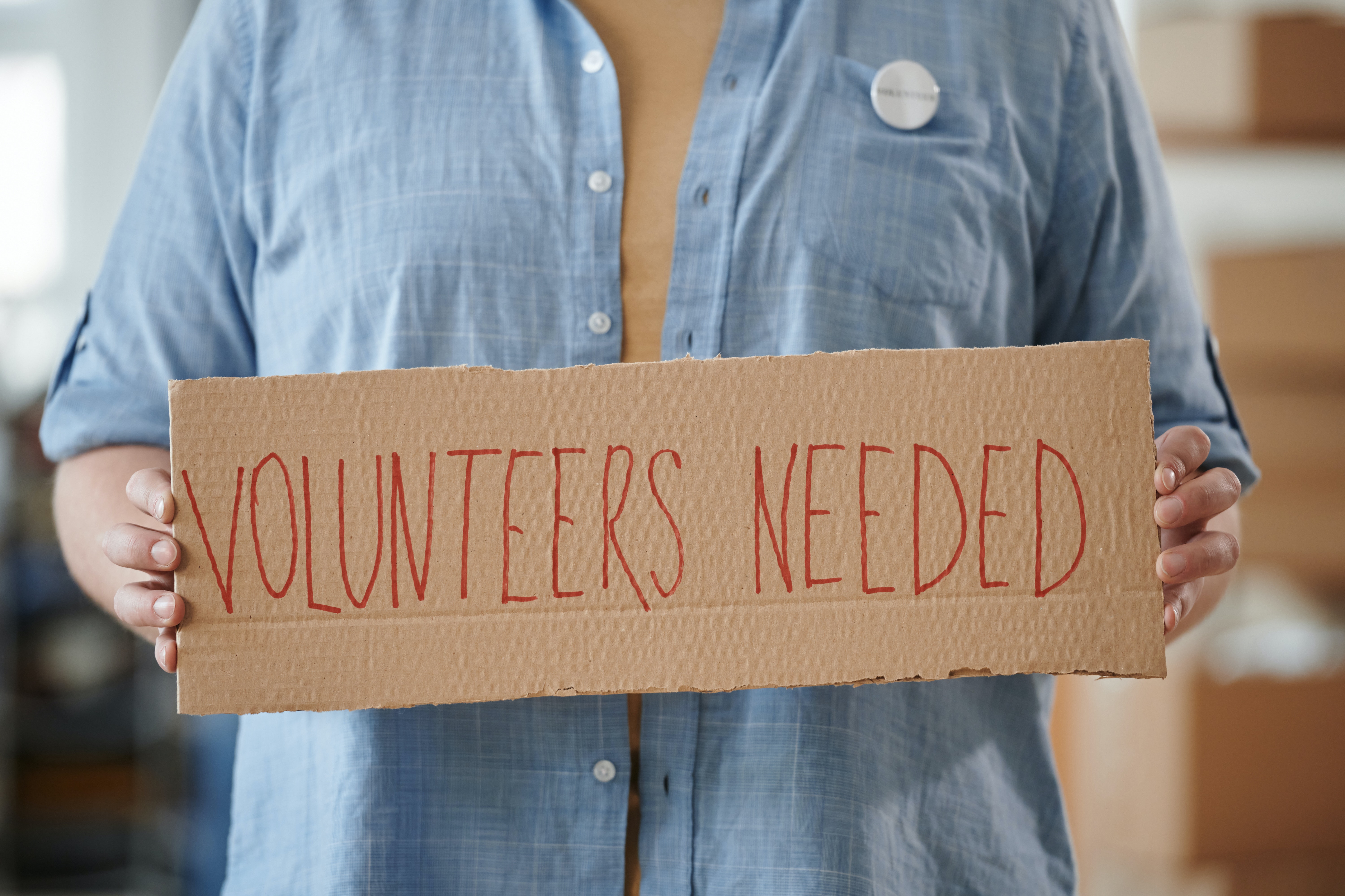 Person holds a cardboard sign saying \"volunteers needed.\"