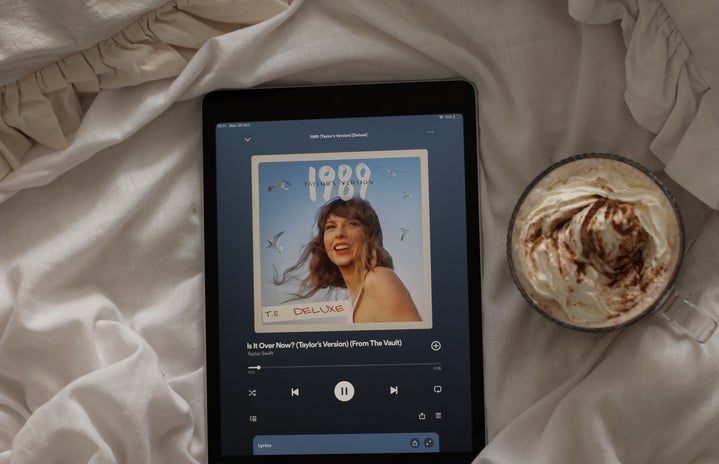 An iPad playing \"Is It Over Now? (Taylor\'s Version) (From The Vault)\" by Taylor Swift on Spotify lying on the white bed sheets next to a mug of hot chocolate
