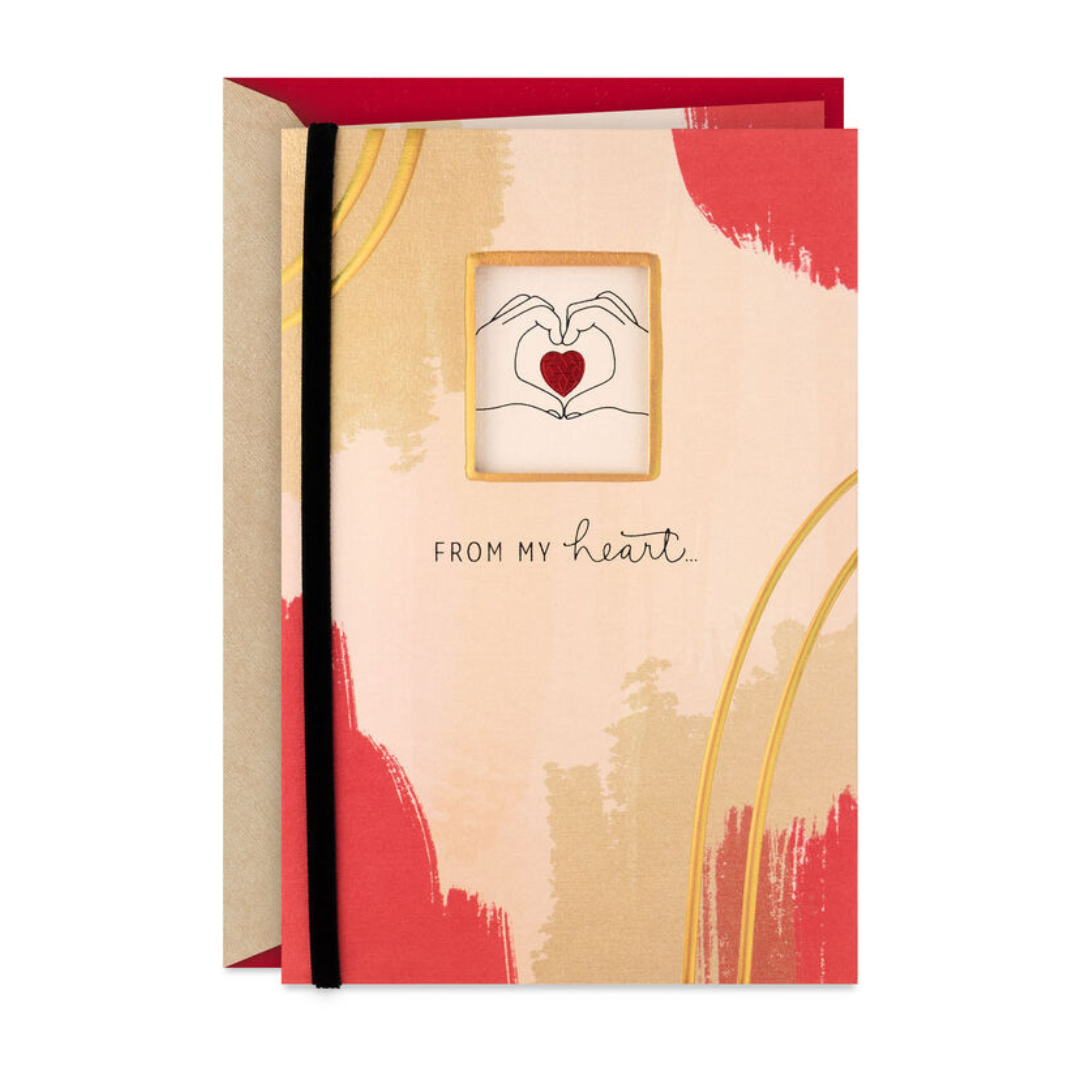 A greeting card with a red and gold abstract design features a small window showing hands forming a heart shape around a red heart. The text reads, \