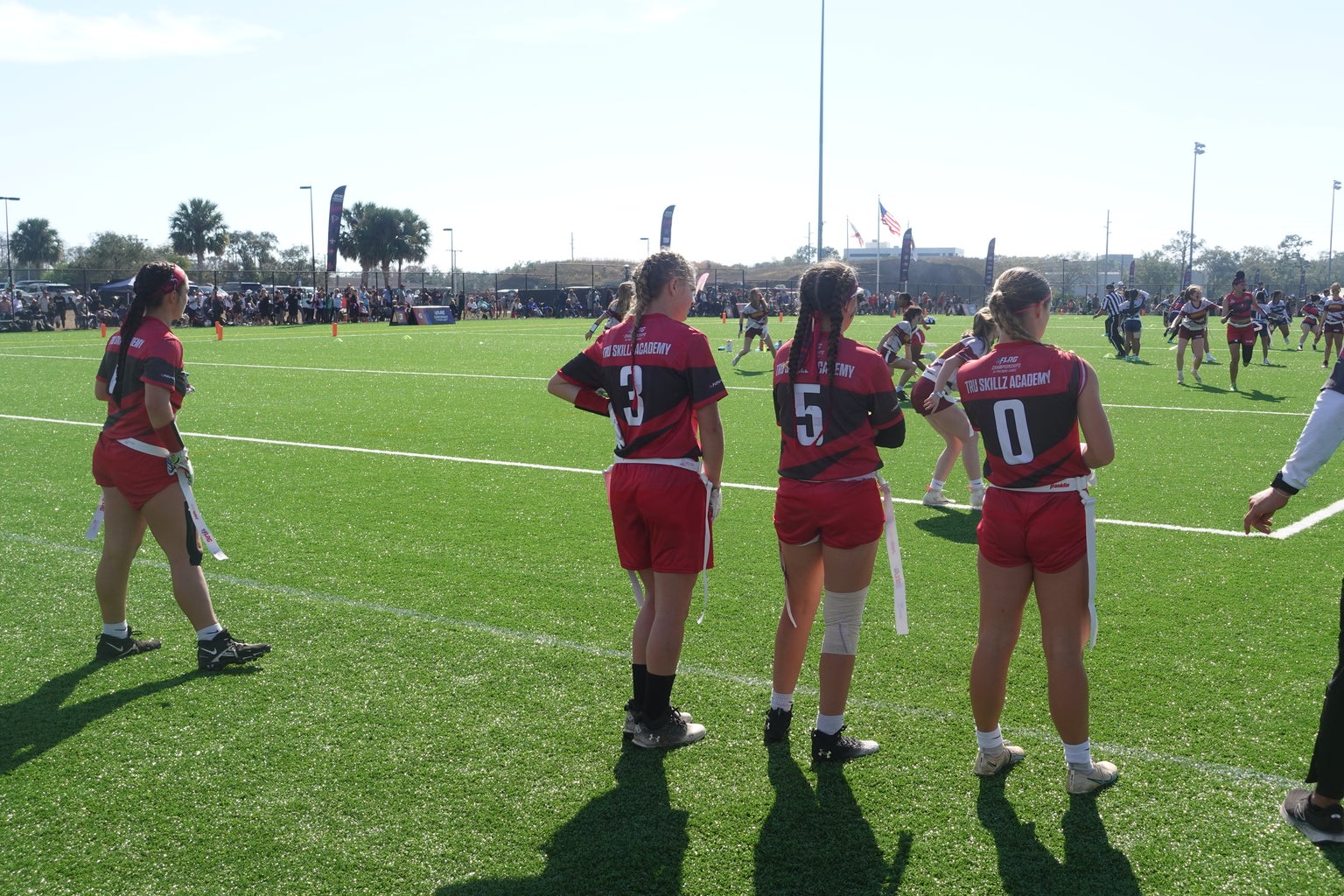 A photo of 4 female flag football players standing on the sideline there is one on the far left and then there are three standing near each other wearing the numbers 3, 5, and 10 all wearing red jerseys