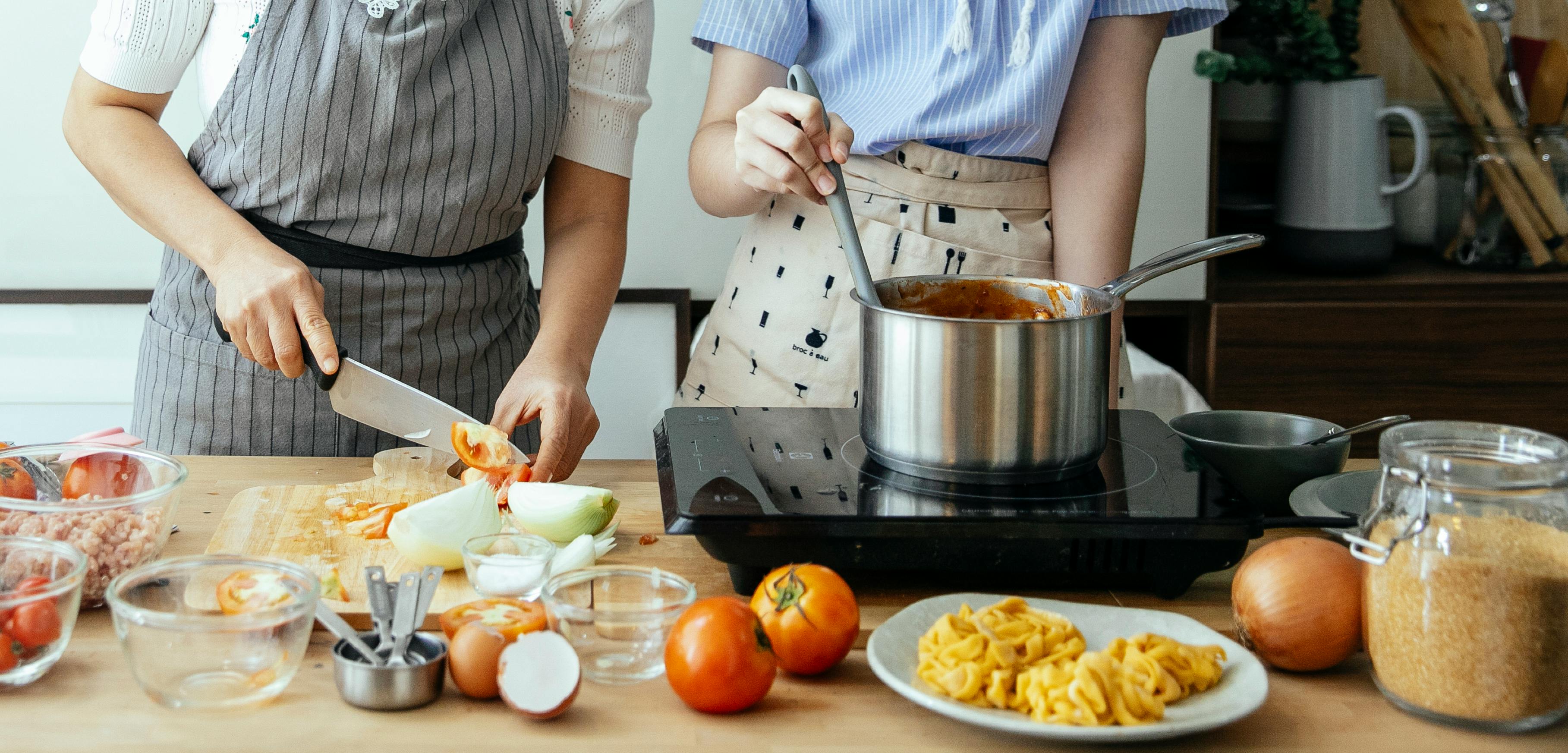 Two women cooking tomato sauce with ingredients laid out.