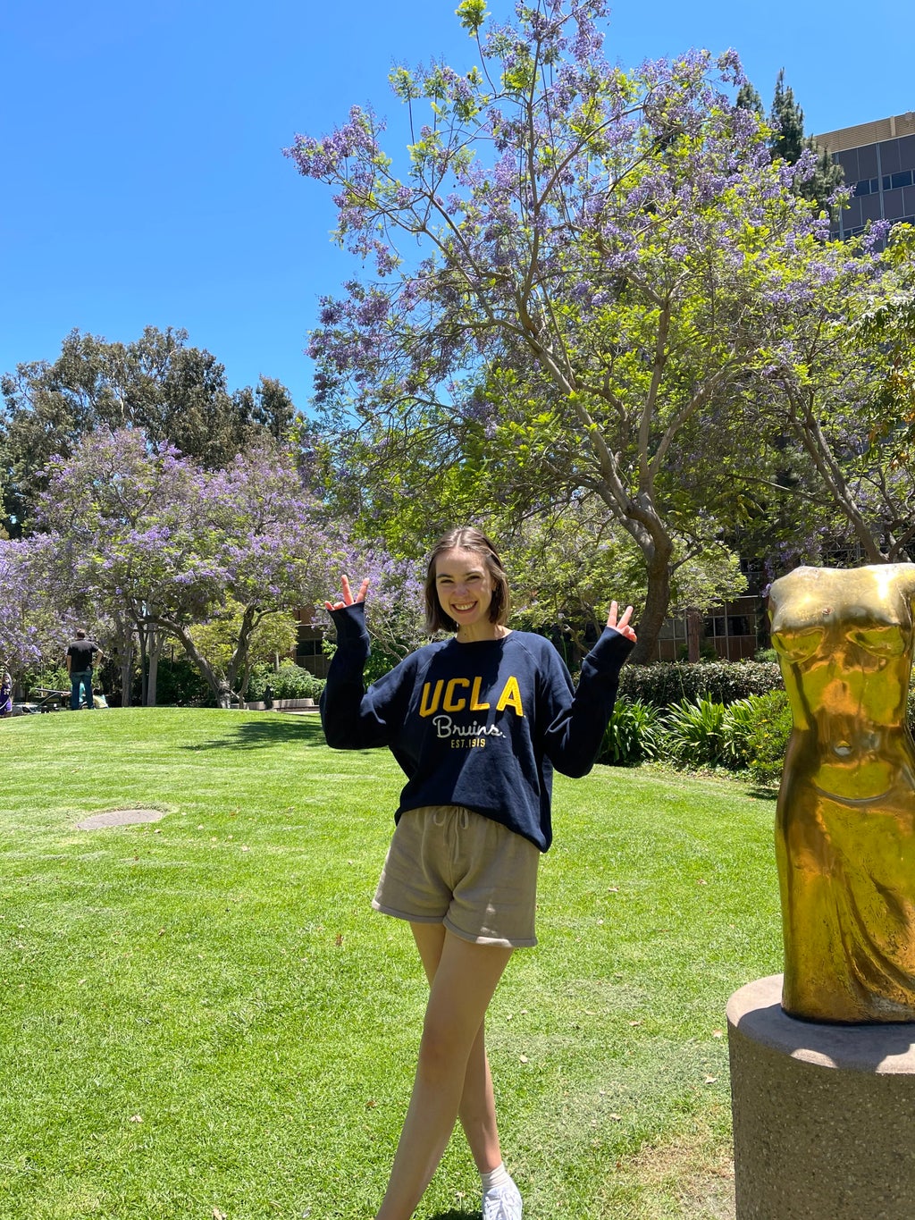 A girl stands in a UCLA sweater in a sunny sculpture garden.