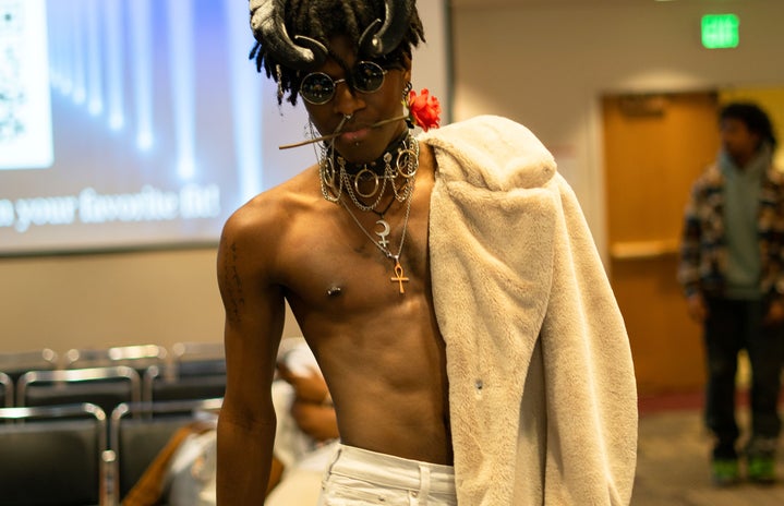Man in fashion show with jacket draped on shoulder, wearing jewelry and head decorations a fashion show