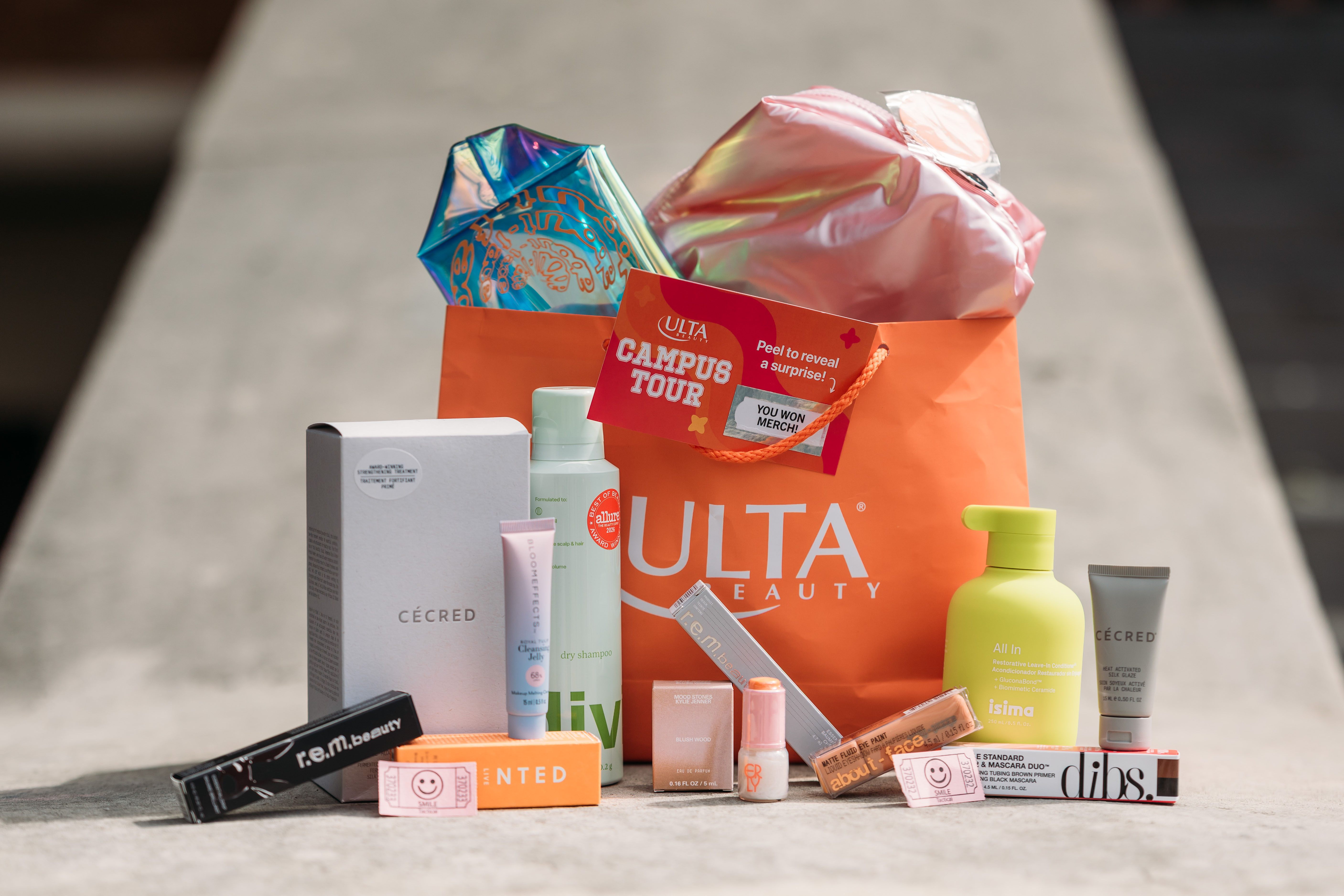 A bright Ulta Beauty shopping bag spills out colorful skincare, makeup and haircare products, coupons, and gift packaging on a stone surface, creating a cheerful haul display.