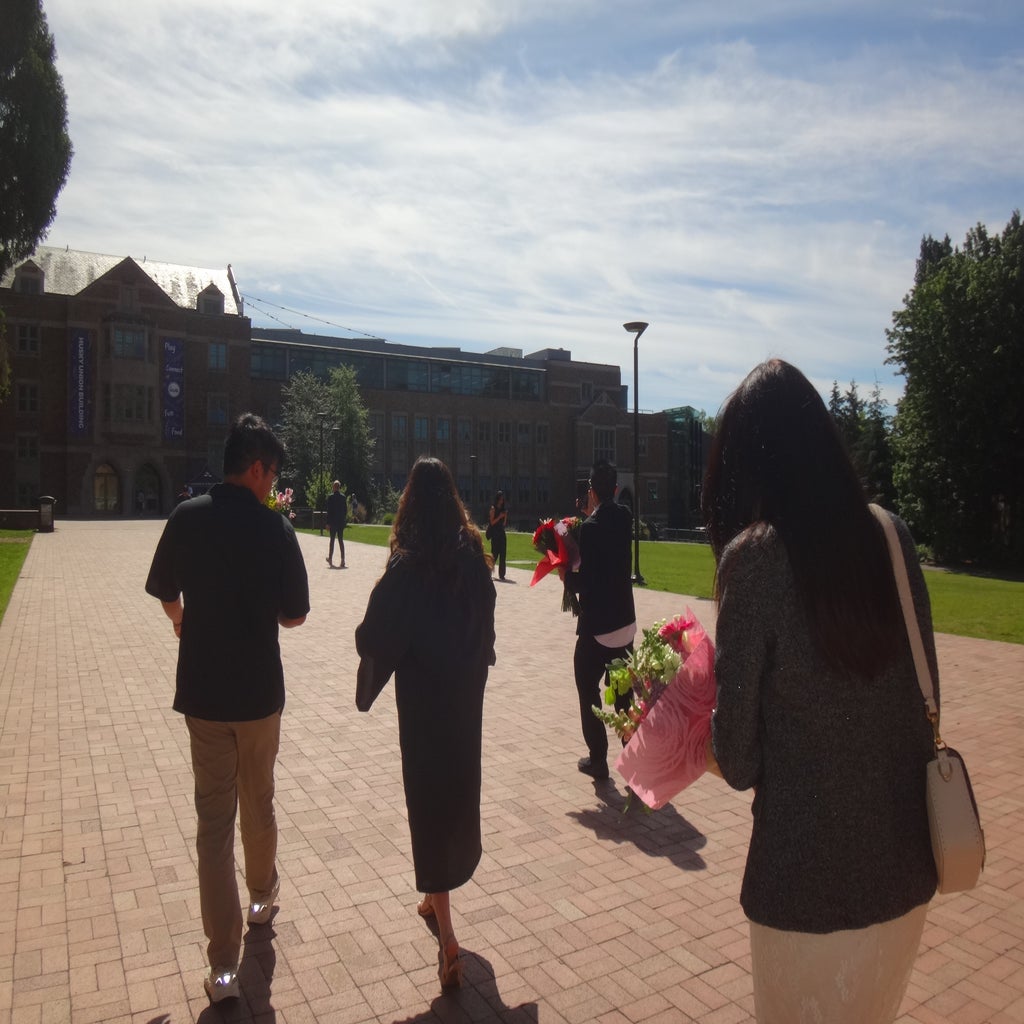 The image shows a group of people walking across a brick plaza on a sunny day, with some carrying bouquets, after graduation.