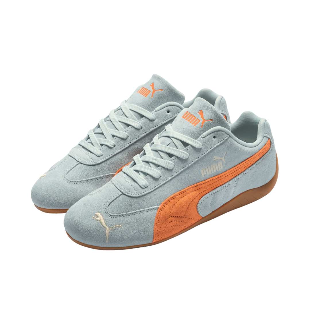 Light gray suede sneakers with orange accents and logos, featuring a sleek design. Sports brand style on a white background. Casual and modern vibe.