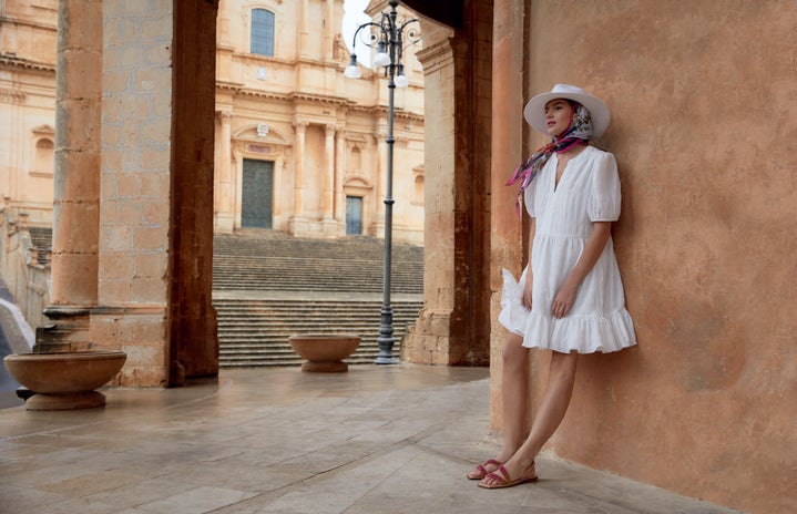 Woman wearing a white linen dress and white hat leaning against a wall