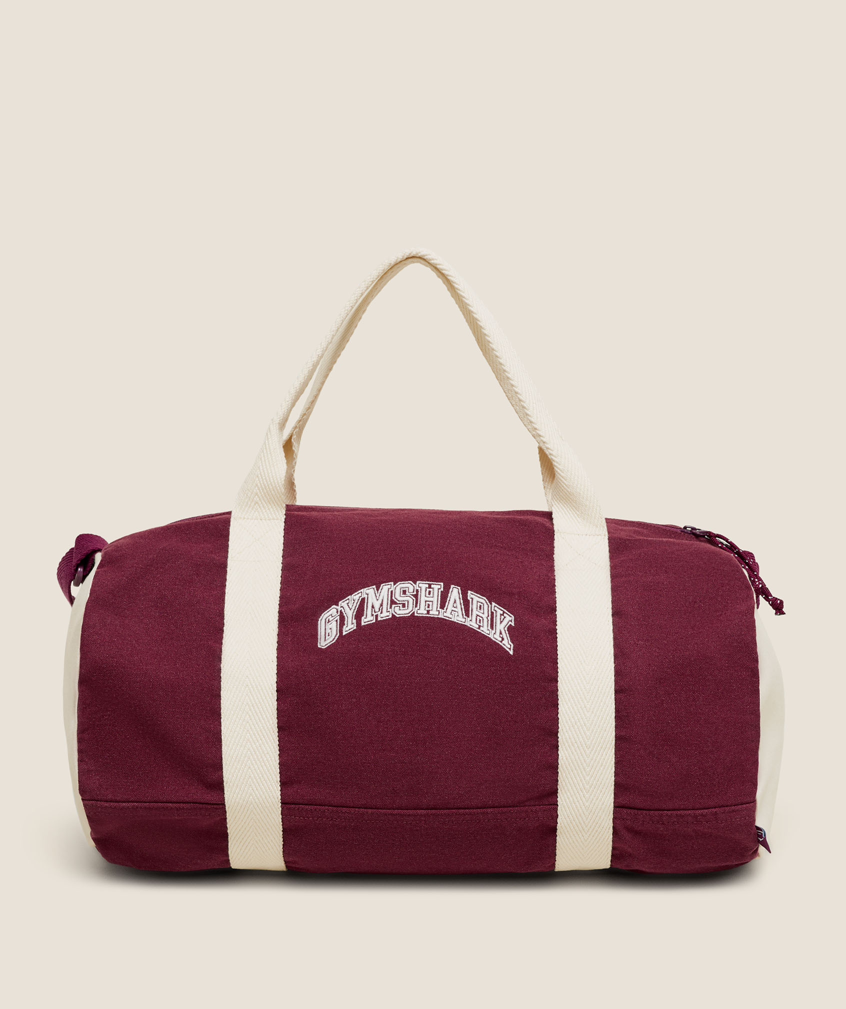 A burgundy Gymshark duffel bag with cream shoulder straps and white logo sits against a plain beige background, conveying a clean, sporty style.