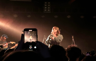 Woman standing on a stage and singing in orange light. A phone films her from the audience.