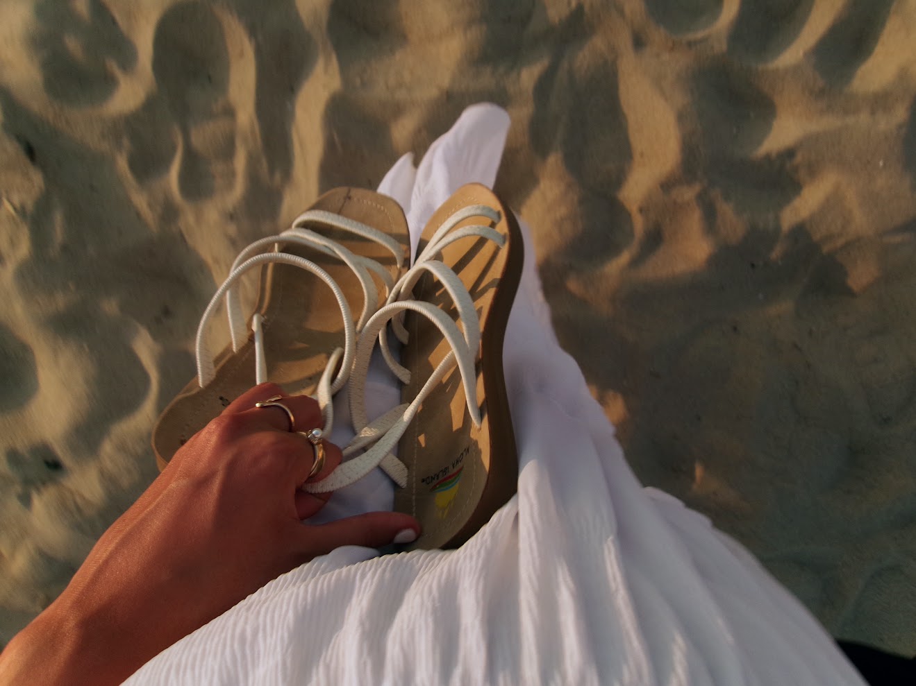 Woman holding sandals on the beach with a white skirt.