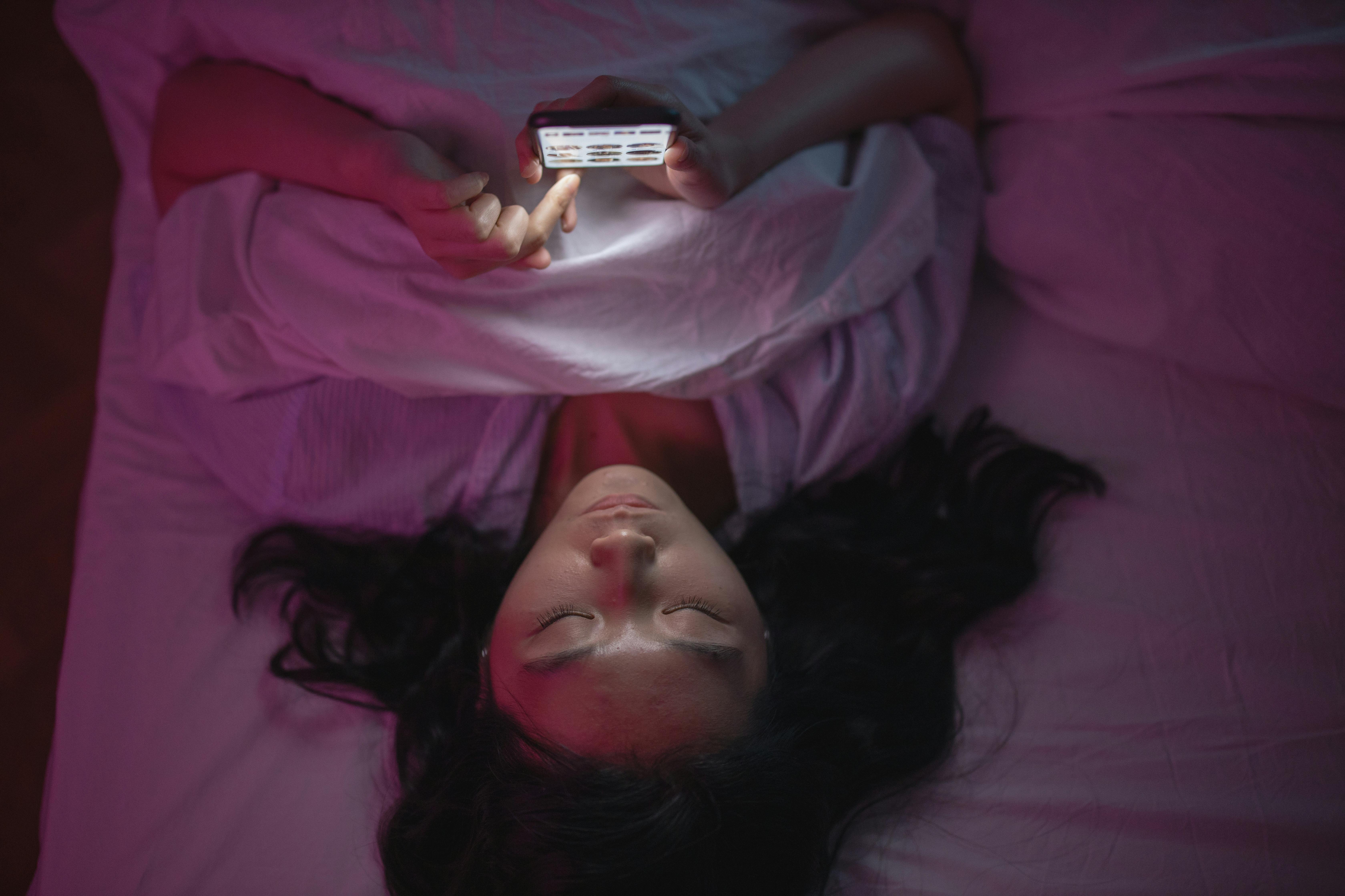 women laying on her back on her bed scrolling on her phone under purple lights