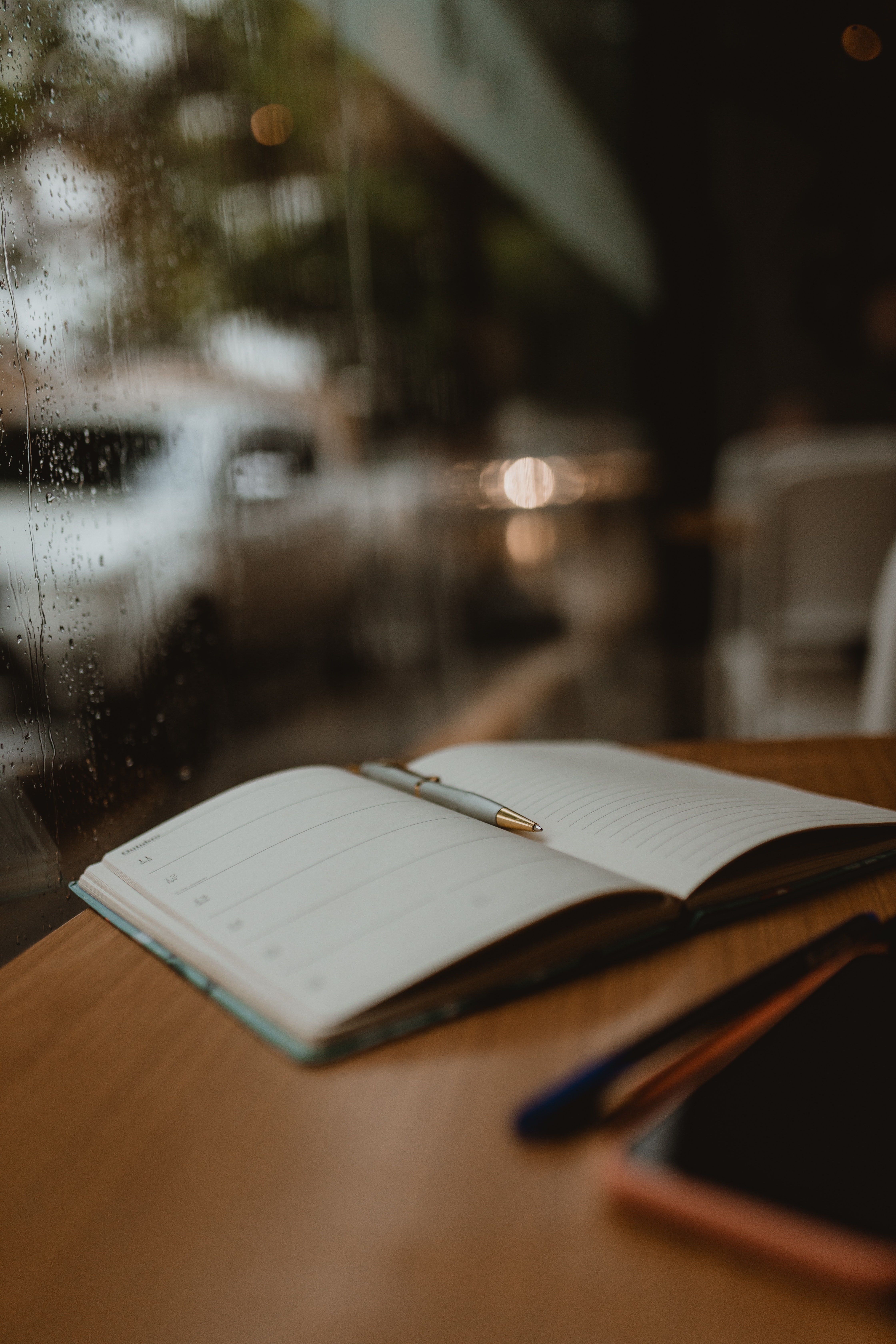 open notebook and pen sit on a table beside a window on a rainy day